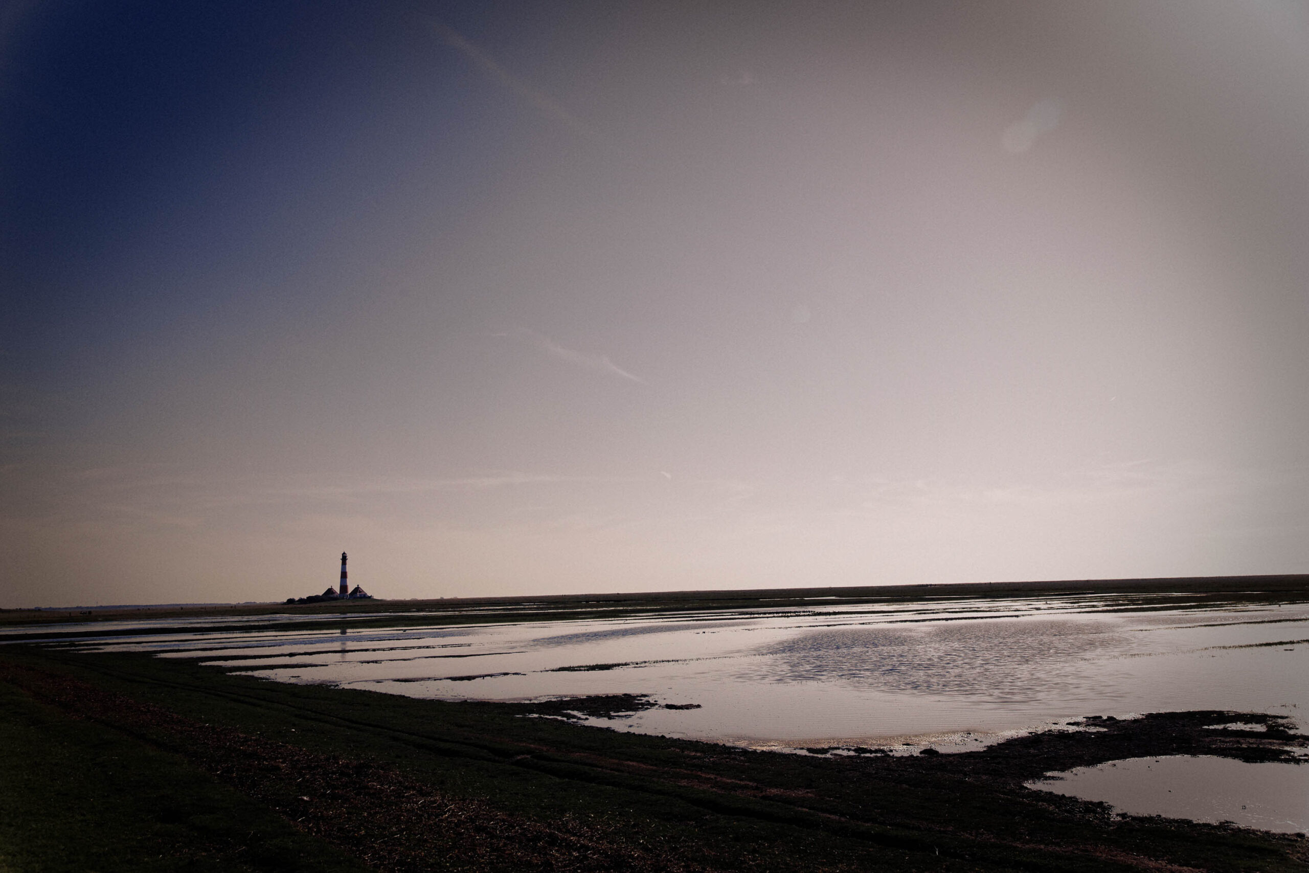 Das Foto zeigt eine düstere Schwarz-Weiß-Szene einer Küstenlandschaft. Im Vordergrund erstreckt sich eine dunkle, sandige Küstenlinie, die von flachem Wasser bedeckt ist, das die schwache Beleuchtung des Himmels reflektiert. Im Hintergrund, silhouettiert gegen den dunklen Himmel, steht der Westerhever Leuchtturm. Der Himmel ist gleichmäßig dunkel, ohne erkennbare Wolken oder Sterne. Die Szene vermittelt eine Atmosphäre der Ruhe und Melancholie.