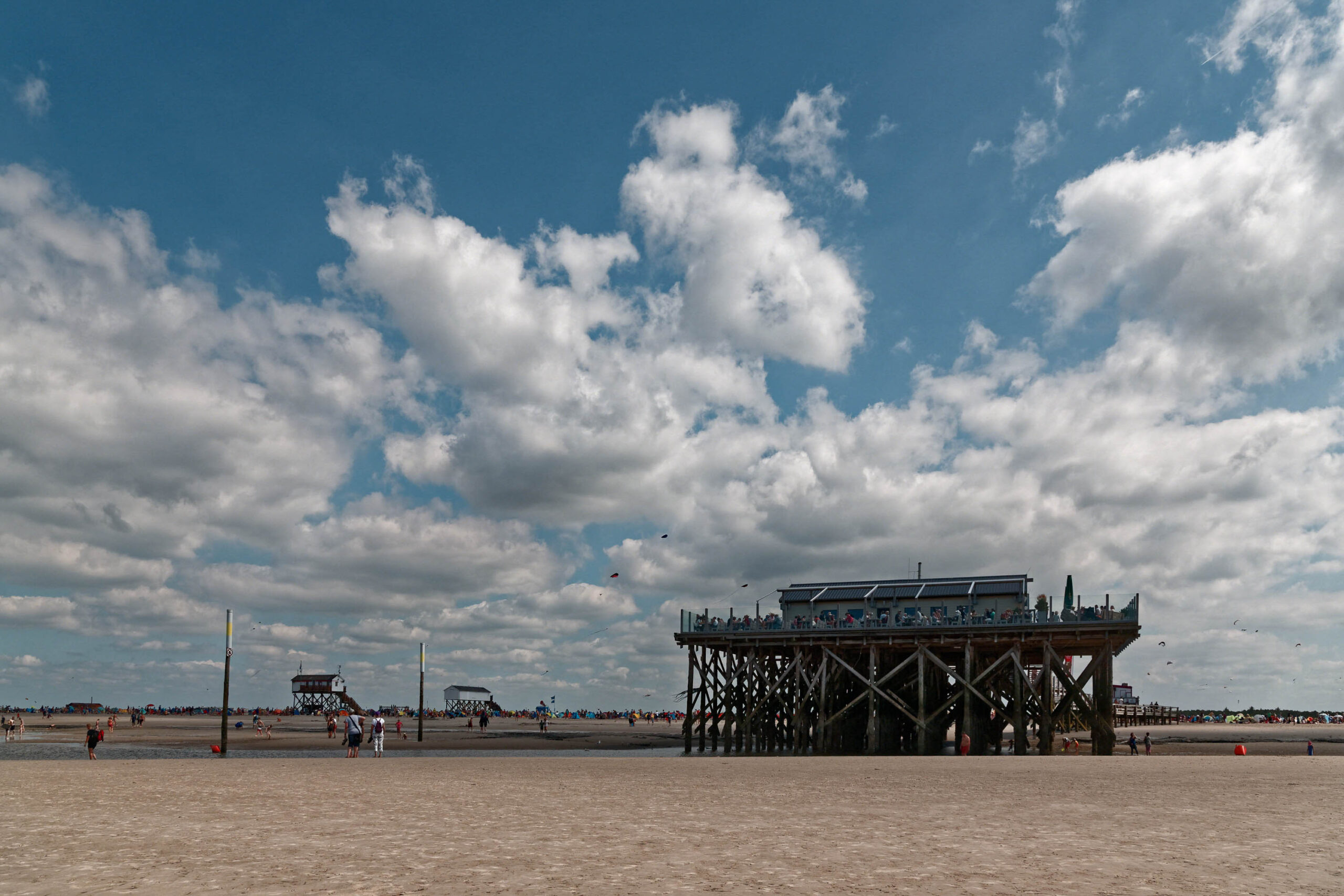 Das Bild zeigt einen Pfahlbau auf einem weitläufigen Sandstrand. Der Himmel ist bewölkt, aber die Sonne scheint durch die Wolken hindurch. Im Hintergrund sind Menschen zu sehen, die sich auf dem Strand aufhalten. Die Szene vermittelt eine sommerliche und entspannte Atmosphäre.