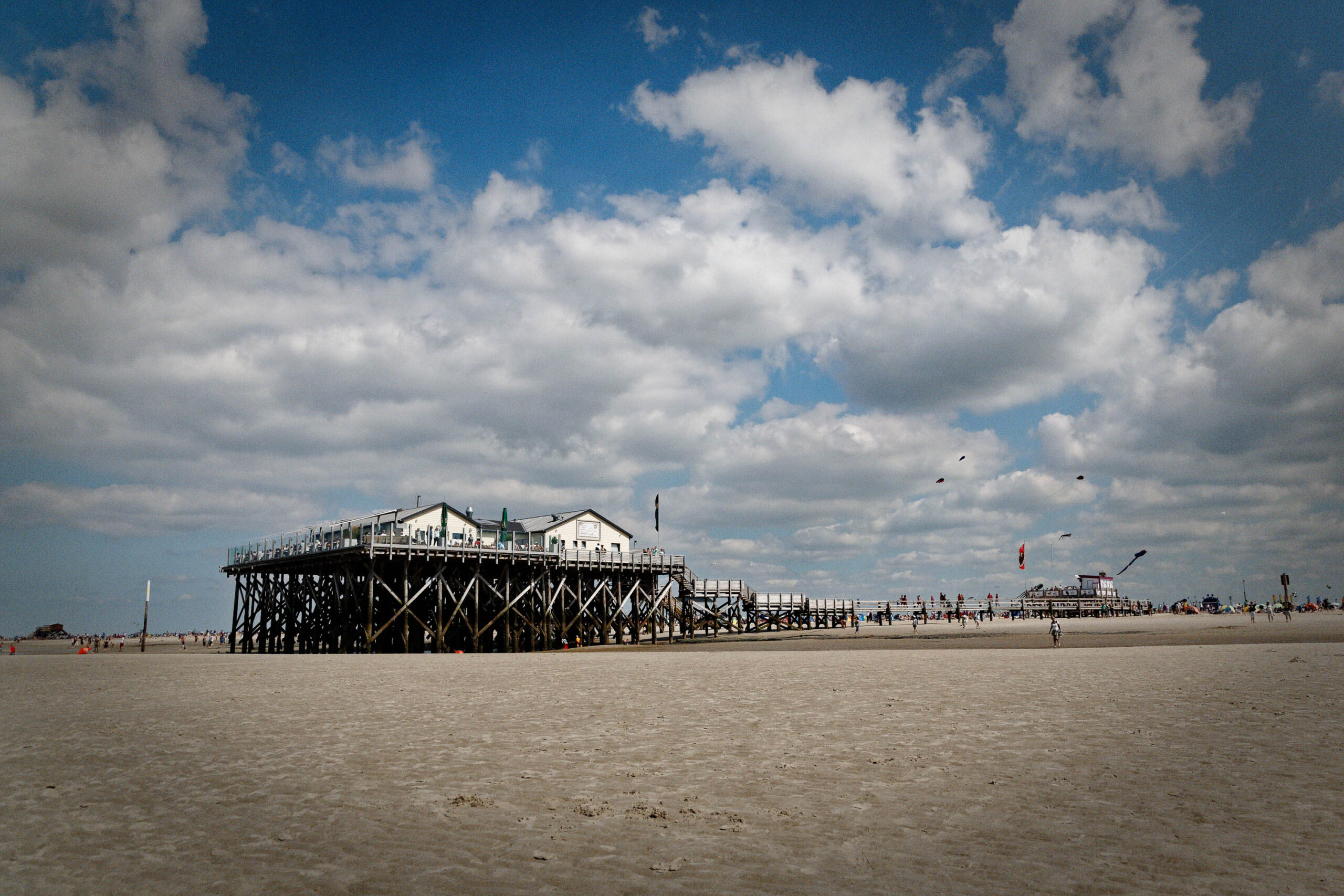 Die Aufnahme zeigt eine Szene am Ordinger Strand in St. Peter-Ording, Deutschland. Im Vordergrund erstreckt sich ein breiter, hellfarbener Sandstrand. Im Mittelpunkt steht ein imposanter Pfahlbau, der auf Stelzen in den Sand gebaut wurde. Der Bau hat mehrere Stockwerke und ist mit Fenstern und Balkonen versehen. Im Hintergrund sind weitere Pfahlbauten und Personen zu sehen, die sich auf dem Strand aufhalten. Der Himmel ist bewölkt, was der Szene eine leicht düstere Atmosphäre verleiht. Die Aufnahme fängt die typische Landschaft einer norddeutschen Küstenregion ein.