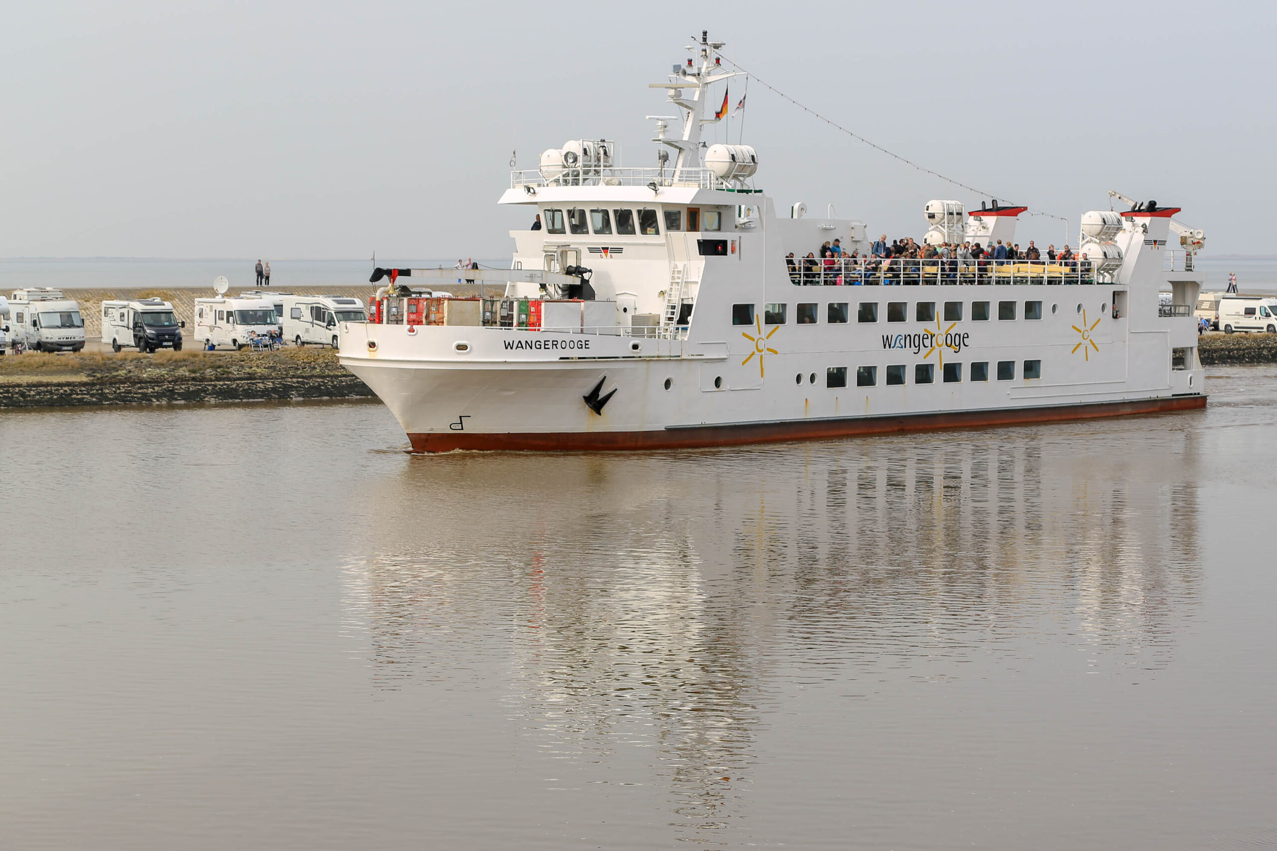 Das Bild zeigt die Fähre 'Wangerooge' beim Einlaufen in einen Hafen. Die Fähre ist weiß und hat mehrere Decks mit Fenstern und Geländern. Auf dem oberen Deck befinden sich Personen. Im Hintergrund sind Wohnmobile und andere Schiffe zu sehen. Der Himmel ist grau und das Wasser ist trüb. Das Bild wurde im April 2018 auf Wangerooge, einer Insel in der Nordsee, aufgenommen.