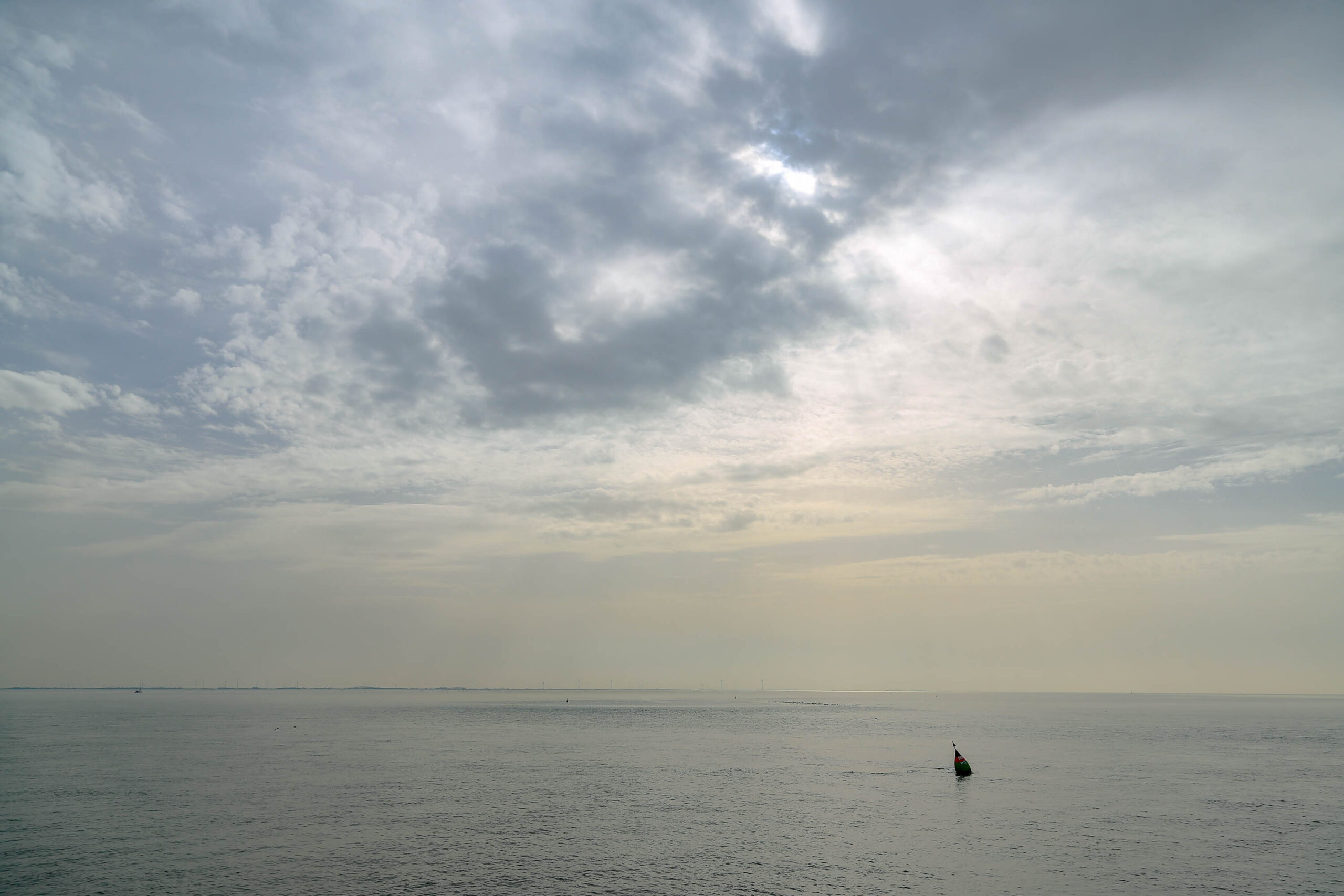 Das Foto zeigt eine ruhige Meereslandschaft, aufgenommen auf der Insel Wangerooge im April 2018. Der Horizont ist kaum erkennbar, da er sich mit dem Himmel vermischt. Eine einzelne rote Boje schwimmt im Wasser, die den Blickfang des Bildes darstellt. Der Himmel ist bedeckt und von Wolken bedeckt, was eine ruhige und friedliche Atmosphäre schafft. Das Licht ist diffus und erzeugt eine sanfte Stimmung.