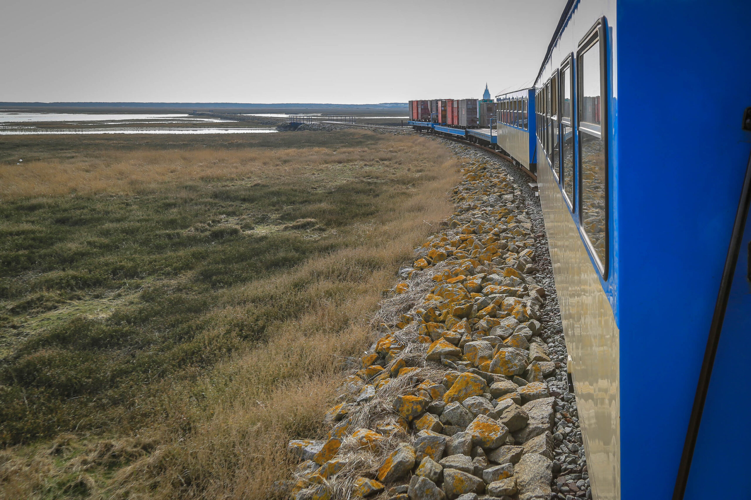 Die Aufnahme zeigt eine Szene von der Insel Wangerooge, Nordsee, im April 2018. Der Blickfang ist ein blau lackierter Zug, der sich auf Schienen durch eine flache, grüne Landschaft bewegt. Links vom Zug erstreckt sich eine weite Ebene aus Gras und einem See oder einer Binnengewässerlandschaft. Die Schienen sind von einem Damm aus Steinen und Felsen flankiert, die mit gelb-orangefarbenem Moos bedeckt sind. Der Himmel ist bedeckt und lässt auf trübes Wetter schließen. Die Perspektive ist aus dem Zugfenster aufgenommen, was den Eindruck einer Reise vermittelt. Die Szene vermittelt eine ruhige und friedliche Atmosphäre, typisch für die norddeutschen Küstenlandschaften.