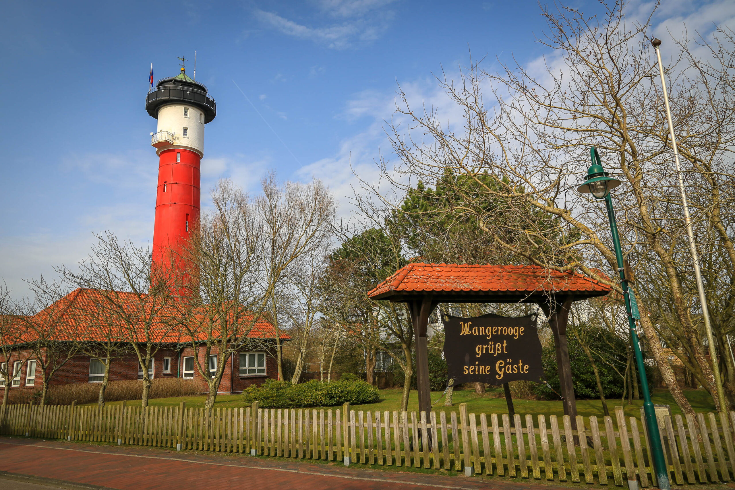 Das Foto zeigt den Wangerooger Leuchtturm, ein markantes Wahrzeichen der Insel Wangerooge. Der Leuchtturm ist rot gestrichen und hat eine weiße Laterne. Er steht auf einem kleinen Hügel, umgeben von Bäumen und einem roten Backsteinhaus. Im Vordergrund befindet sich ein hölzernes Schild mit der Aufschrift 'Wangerooge grüßt seine Gäste'. Der Himmel ist blau und wolkenlos, was auf einen sonnigen Tag hindeutet. Die Szene vermittelt eine friedliche und einladende Atmosphäre.