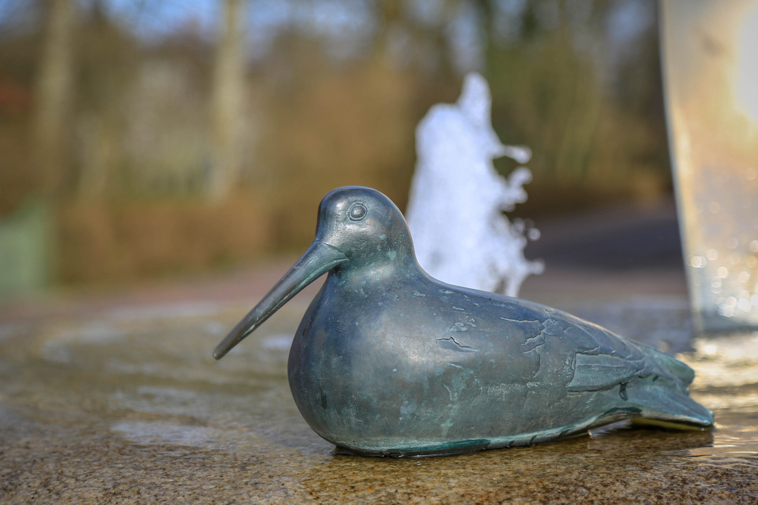 Das Bild zeigt eine bronzene Vogelstatue, die in einem flachen Wasserbecken sitzt. Der Vogel ist dunkelgrün und hat einen langen, spitzen Schnabel. Im Hintergrund ist ein Springbrunnen zu sehen, der Wasser in die Luft sprüht. Das Bild ist in einem warmen Licht gehalten, was eine ruhige und friedliche Atmosphäre schafft. Der Hintergrund ist unscharf, was den Fokus auf die Vogelstatue lenkt. Die Aufnahme wurde vermutlich im April 2018 auf der Insel Wangerooge in der Nordsee aufgenommen.