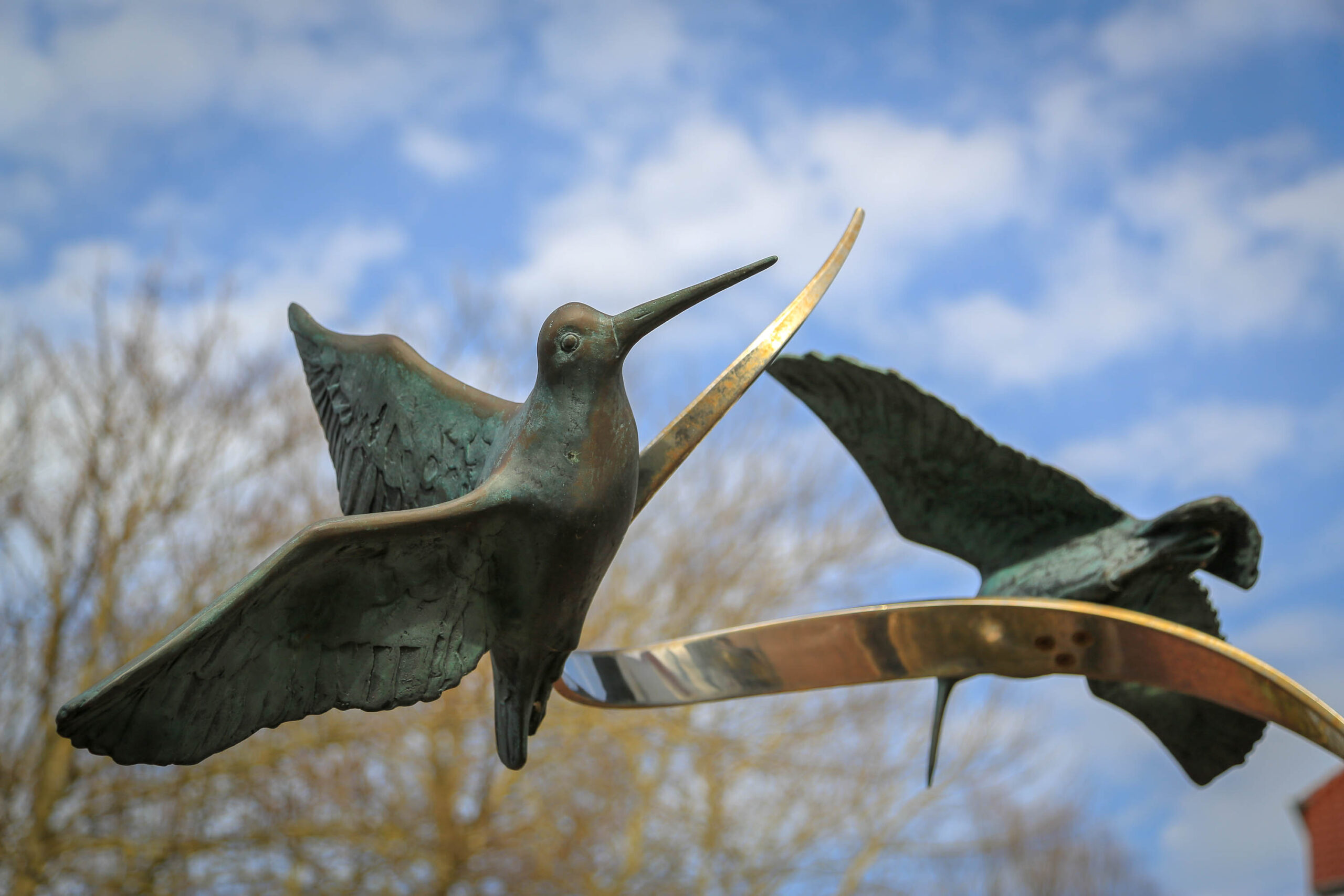Das Bild zeigt eine Bronzestatue von zwei Seeschwalben, die in der Flugposition dargestellt sind. Die Vögel sitzen auf einem geschwungenen, goldenen Metallbogen, der an einen stilisierten Wellenreiter erinnert. Der Hintergrund besteht aus einem blauen Himmel mit vereinzelten weißen Wolken und einem Hauch von braunen Ästen. Die Statue ist detailliert gearbeitet und fängt die Dynamik des Flugs der Seeschwalben gut ein. Die Beleuchtung ist weich und gleichmäßig, was die Farben der Statue und des Hintergrunds hervorhebt. Die Komposition ist ausgewogen und harmonisch, wobei die Statue im Mittelpunkt steht und der Himmel als Kontrast dient.
