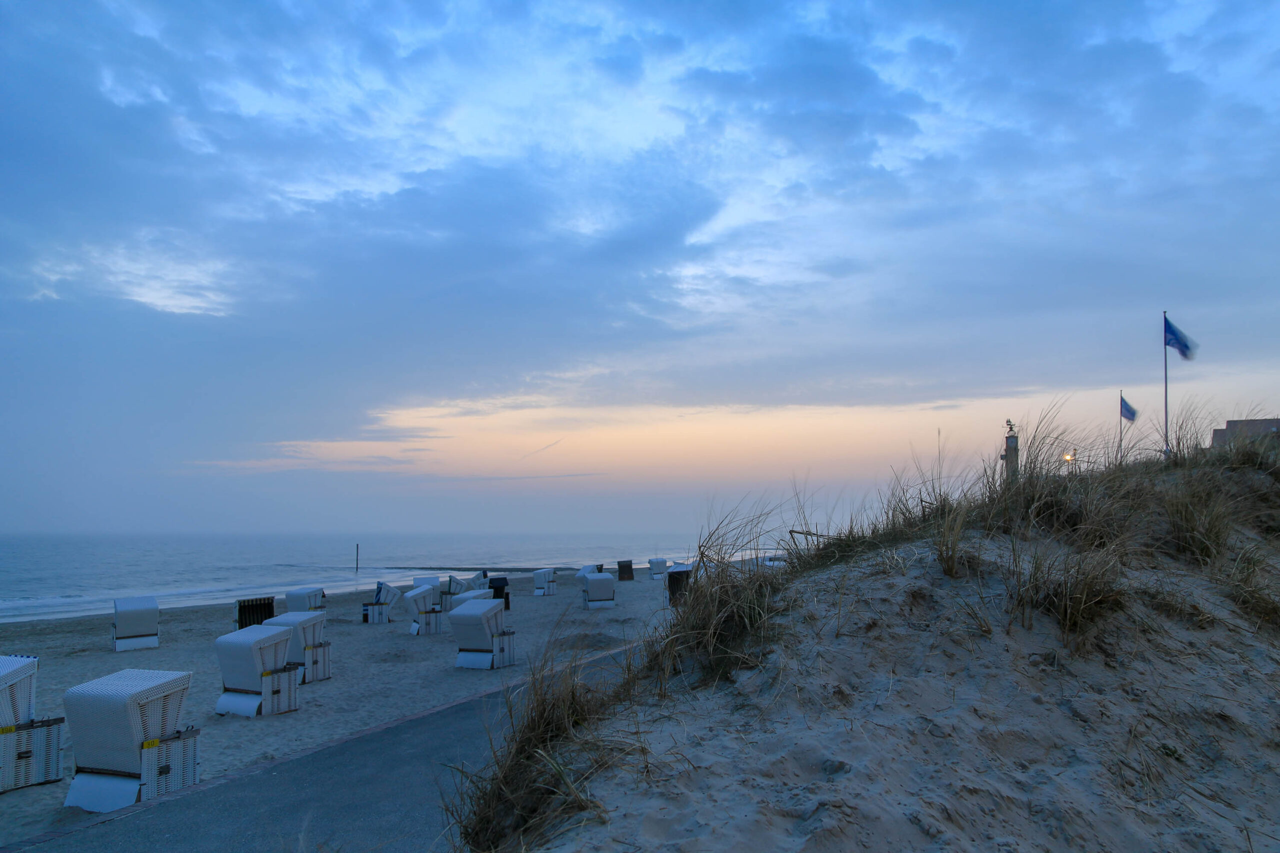 Das Bild zeigt eine ruhige Strandszene auf der Insel Wangerooge, Nordsee. Die Dämmerung hat bereits begonnen, und der Himmel ist in sanften Blautönen und einem Hauch von Orange gefärbt. Im Vordergrund erstrecken sich Reihen von weißen Strandkörben, die für die Erholung der Besucher bereitstehen. Die Dünen, bewachsen mit Gras und einigen wenigen Pflanzen, bilden einen natürlichen Übergang zum Strand. Das Meer ist ruhig und spiegelt die Farben des Himmels wider. Im Hintergrund sind einige Gebäude und ein Leuchtturm erkennbar, die die Insel Wangerooge prägen. Die Szene vermittelt ein Gefühl von Ruhe, Frieden und Erholung.