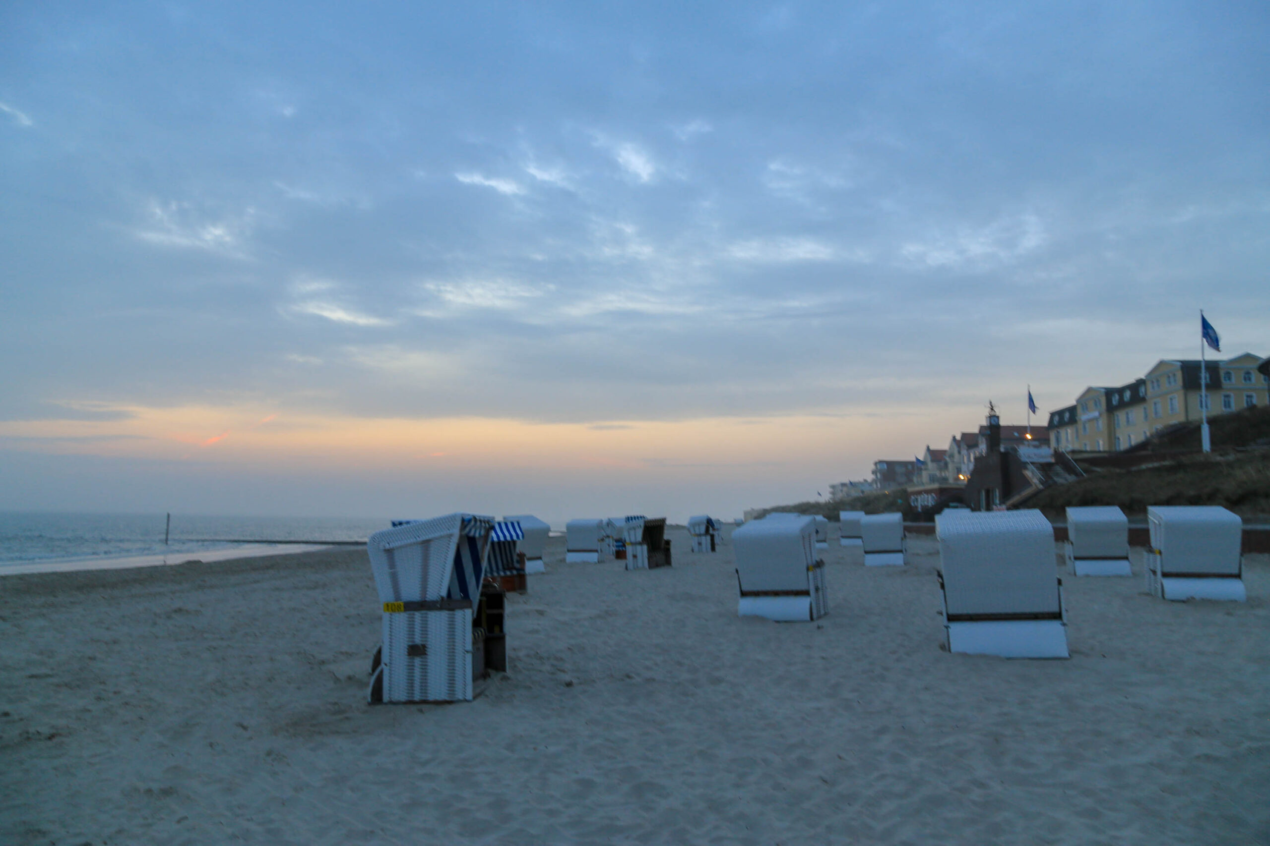 Das Bild zeigt einen Strand bei Sonnenuntergang auf der Insel Wangerooge. Im Vordergrund stehen zahlreiche Strandkörbe, die in einer Reihe aufgereiht sind. Der Strand ist mit feinem Sand bedeckt. Im Hintergrund sind Häuser zu sehen, die sich leicht in den Hang gebaut haben. Der Himmel ist in warmen Farben von Orange und Blau gemischt, was eine ruhige und friedliche Atmosphäre schafft. Die Beleuchtung der Häuser im Hintergrund deutet auf den Beginn der Abendstimmung hin.