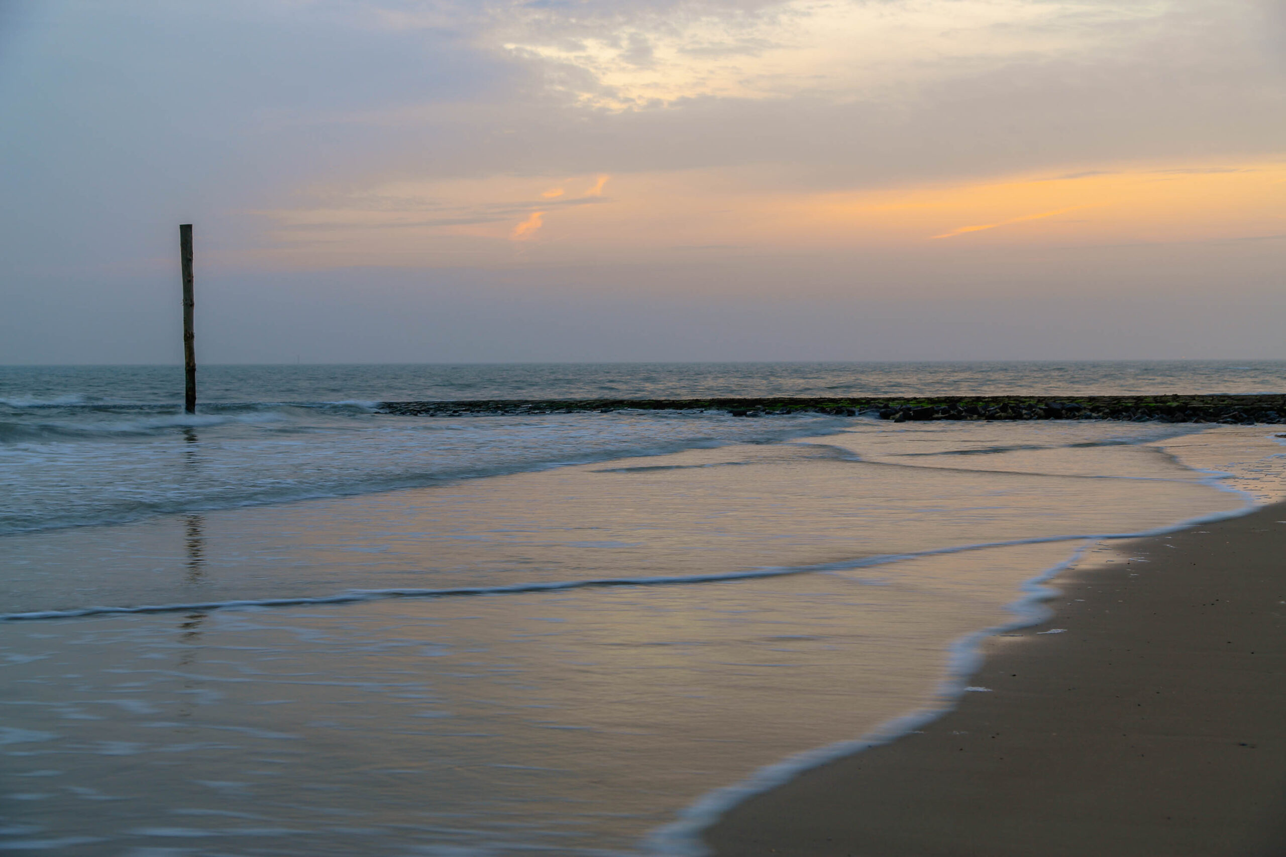 Das Bild zeigt einen Strand bei Sonnenuntergang oder Sonnenaufgang. Der Himmel ist in warmen Farbtönen von Orange und Rosa gehalten, was eine ruhige und friedliche Atmosphäre erzeugt. Der Strand ist mit feinem Sand bedeckt, der vom sanften Wellengang benetzt wird. Im Vordergrund steht ein Holzpfahl, der aus dem Wasser ragt, möglicherweise ein Teil einer alten Schutzstruktur oder ein Meereszeichen. Die Szene vermittelt ein Gefühl von Weite und Stille, typisch für die Nordsee-Küste.