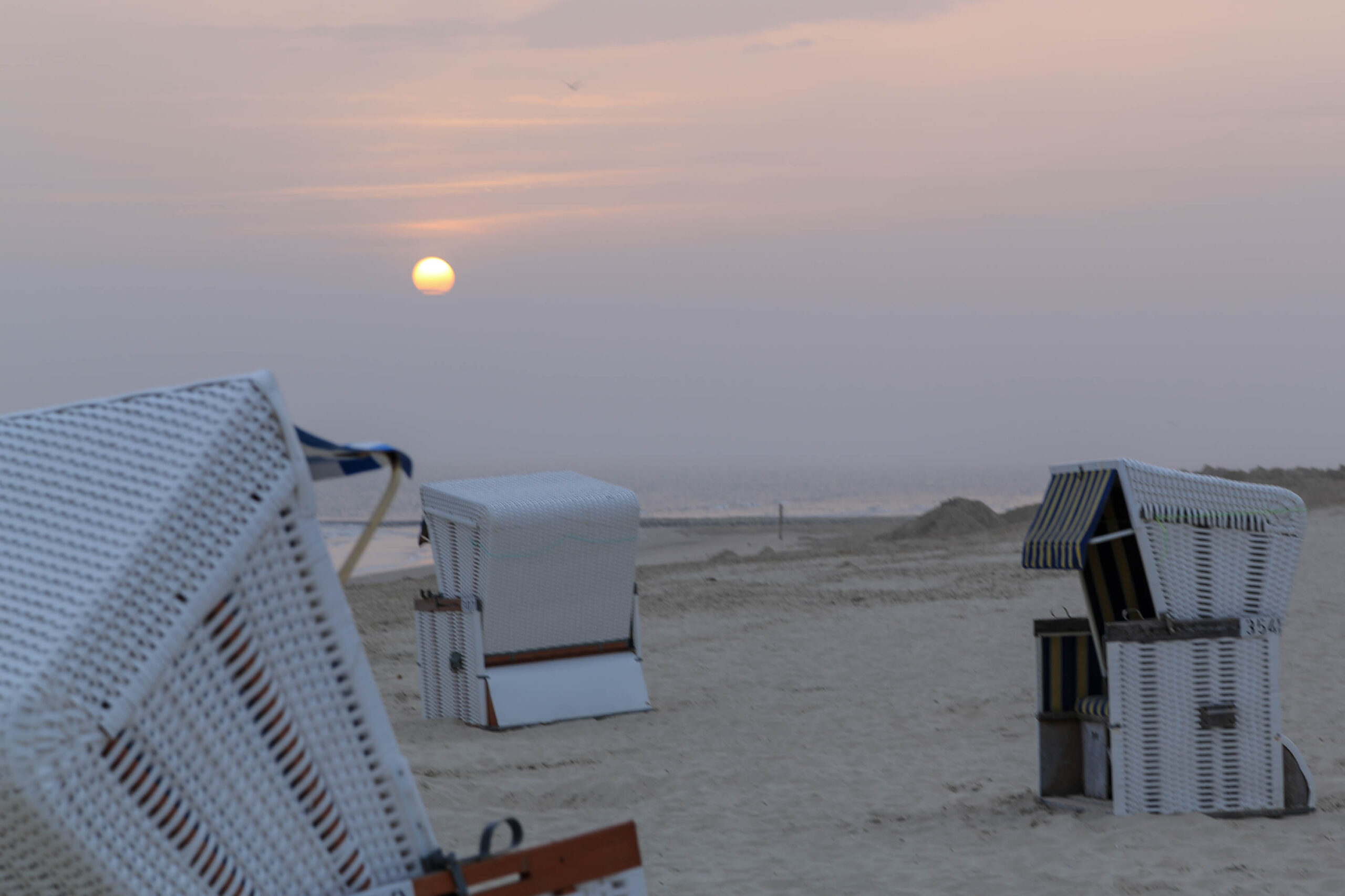 Die Aufnahme zeigt eine friedliche Szene am Strand von Wangerooge, Nordsee. Im Vordergrund stehen mehrere typische Strandkörbe aus Weide, die in einem warmen Licht erstrahlen. Der Himmel ist in sanften Pastelltönen gehalten, was eine ruhige und entspannte Atmosphäre schafft. Der Strand erstreckt sich bis zum Horizont, wo er in einem Dunst verschwimmt. Die Szene vermittelt ein Gefühl von Ruhe und Abgeschiedenheit, typisch für die Nordseeinseln.