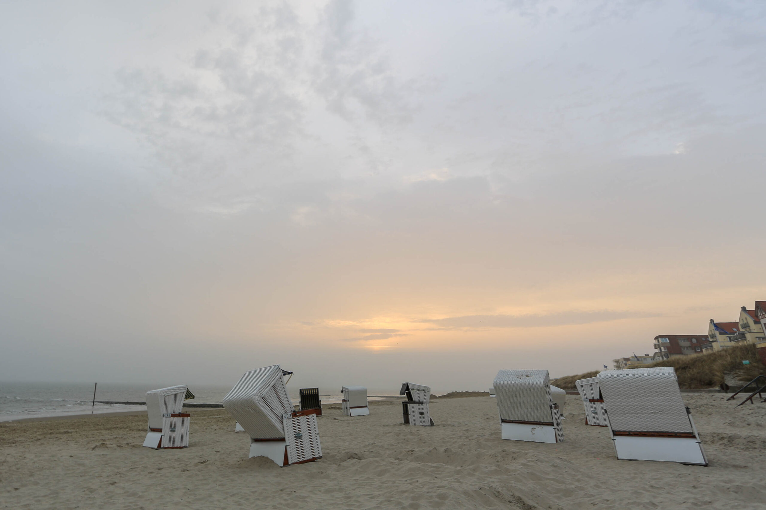 Das Bild zeigt einen Sandstrand bei Sonnenuntergang auf der Insel Wangerooge. Im Vordergrund stehen mehrere weiße Strandkörbe, die in unterschiedlichen Winkeln aufgestellt sind. Der Sand ist hell und feucht. Im Hintergrund ist das Meer zu sehen, das in einen trüben Himmel übergeht. Am Horizont sind einige Gebäude erkennbar. Die Szene vermittelt eine ruhige und friedliche Atmosphäre.