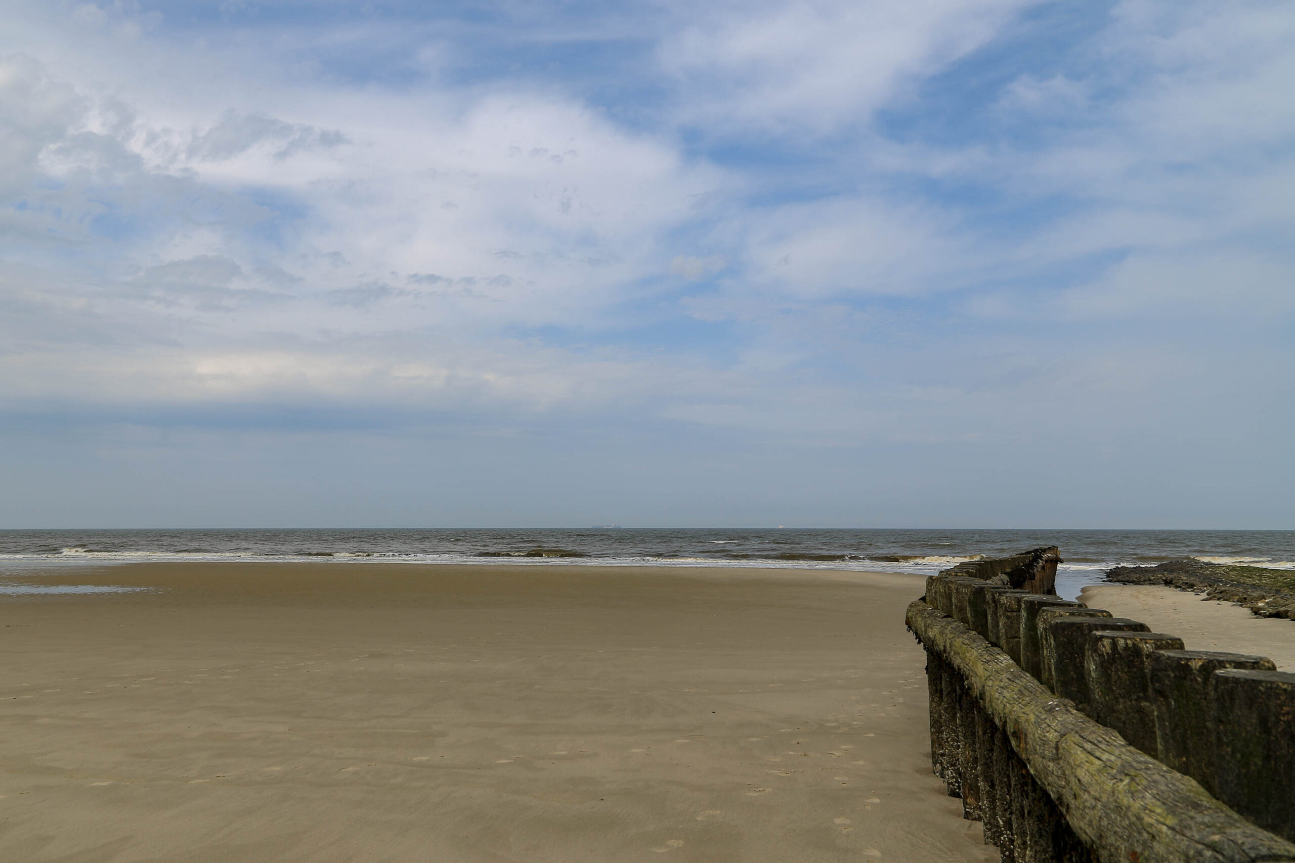 Die Aufnahme zeigt einen typischen Strandabschnitt auf Wangerooge, einer der Ostfriesischen Inseln. Der feine, helle Sand erstreckt sich weitläufig, und im Vordergrund ist ein alter Holzpflockzaun zu sehen, der vermutlich als Schutz vor dem Meer dient. Der Himmel ist bedeckt, was auf ein eher trübes Wetter hindeutet. Der Ozean ist ruhig, aber die Wellen brechen sanft am Strand. Die Perspektive ist weitwinkelig, was die Weite des Strandes betont. Die Aufnahme vermittelt eine ruhige und friedliche Atmosphäre.