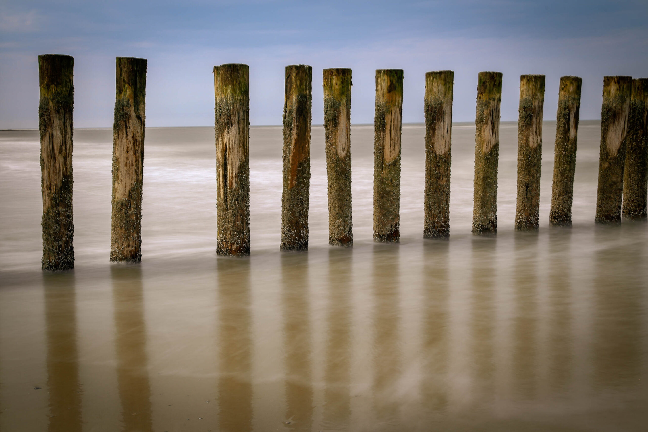 Das Bild zeigt eine Reihe von verwitterten Holzpfählen, die aus dem trüben Wasser der Nordsee ragen. Die lange Belichtungszeit hat dazu geführt, dass das Wasser wie ein milchiger Schleier wirkt und die Bewegung des Wassers betont. Der Himmel ist leicht bewölkt und trägt zur ruhigen Atmosphäre des Bildes bei. Die Pfähle scheinen eine Art Schutzstruktur oder eine alte Mole zu markieren. Die Szene vermittelt ein Gefühl von Vergänglichkeit und die Kraft der Natur.