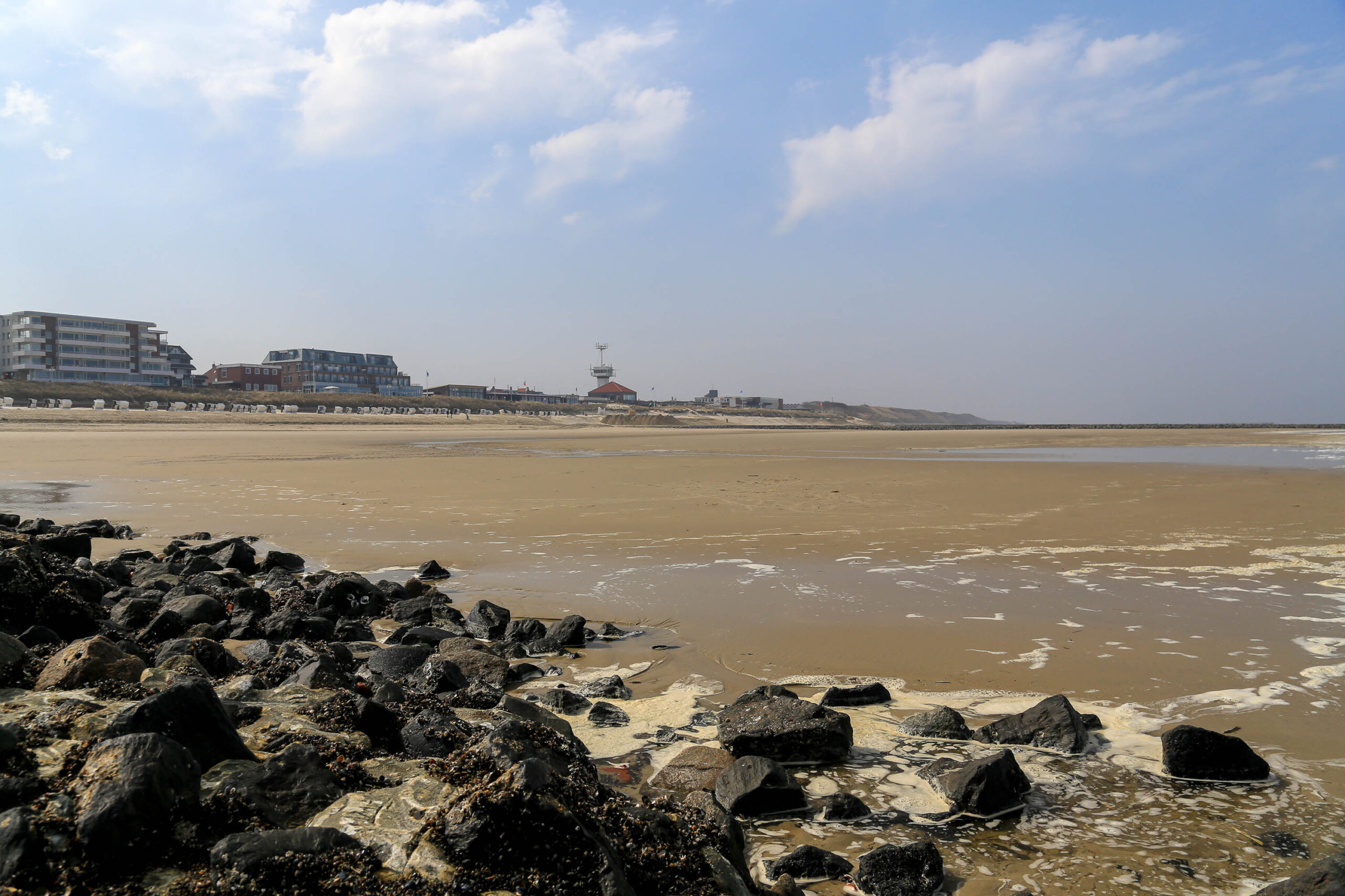 Die Aufnahme zeigt einen weiten Sandstrand auf der Insel Wangerooge. Der Strand ist bei Ebbe freigelegt, und das Wasser steht sehr weit entfernt. Im Hintergrund sind Gebäude zu sehen, die wahrscheinlich Hotels oder Ferienwohnungen sind. Der Himmel ist blau mit vereinzelten weißen Wolken. Die Szene vermittelt eine ruhige und friedliche Atmosphäre. Die Felsen im Vordergrund sind mit Seetang bewachsen.
