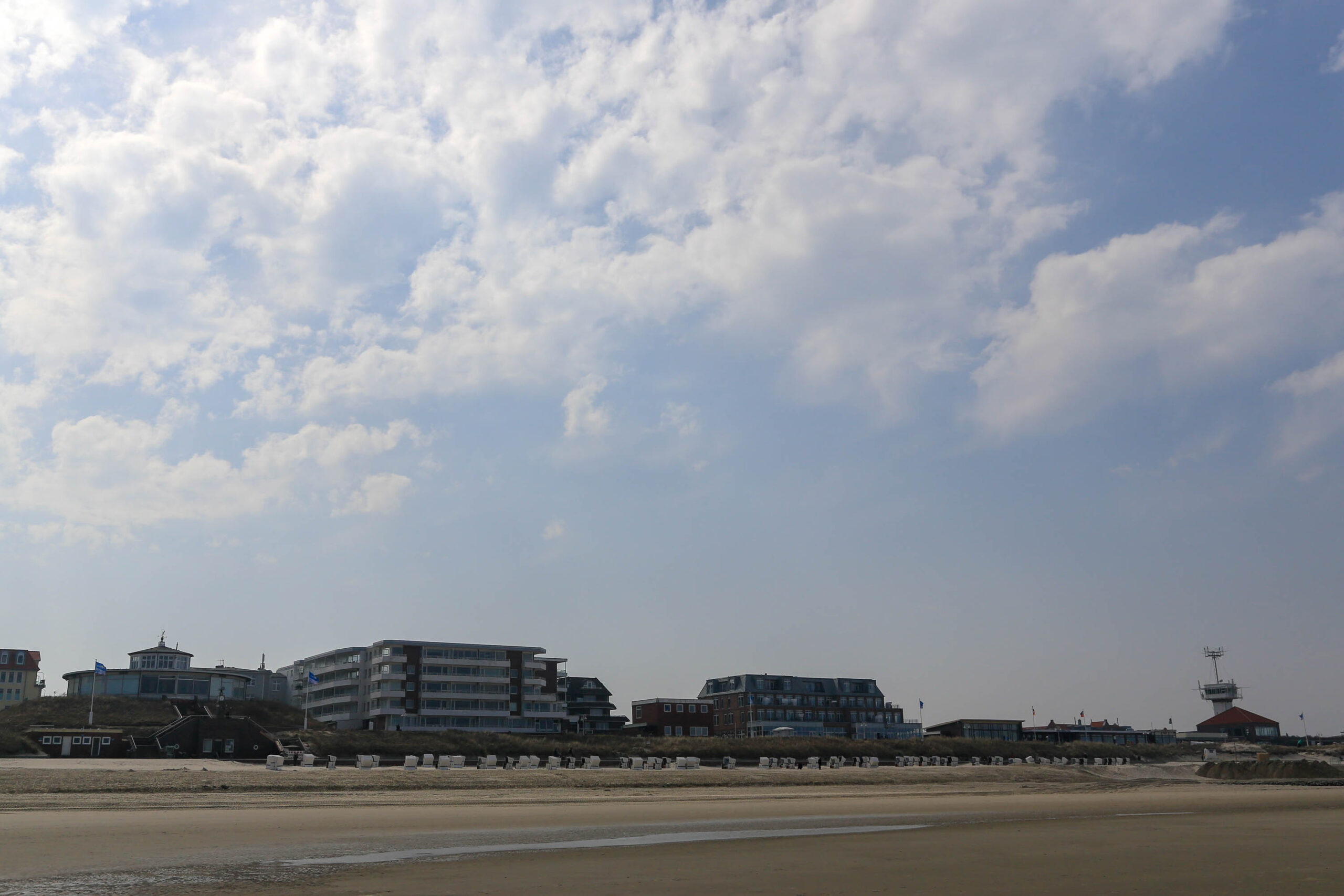 Die Aufnahme zeigt eine typische Szene von Wangerooge, einer Insel in der Deutschen Nordsee. Im Vordergrund erstreckt sich ein breiter Sandstrand, der mit zahlreichen Liegestühlen für Sonnenanbeter bestückt ist. Im Hintergrund sind mehrere Gebäude zu sehen, die wahrscheinlich Hotels oder Ferienwohnungen sind. Die Architektur ist typisch für norddeutsche Küstenorte, mit roten Ziegeldächern und weißen Fassaden. Der Himmel ist von einer Schicht aus Wolken bedeckt, die das Licht streuen und eine sanfte Atmosphäre schaffen. Die Aufnahme wurde im April 18 aufgenommen.