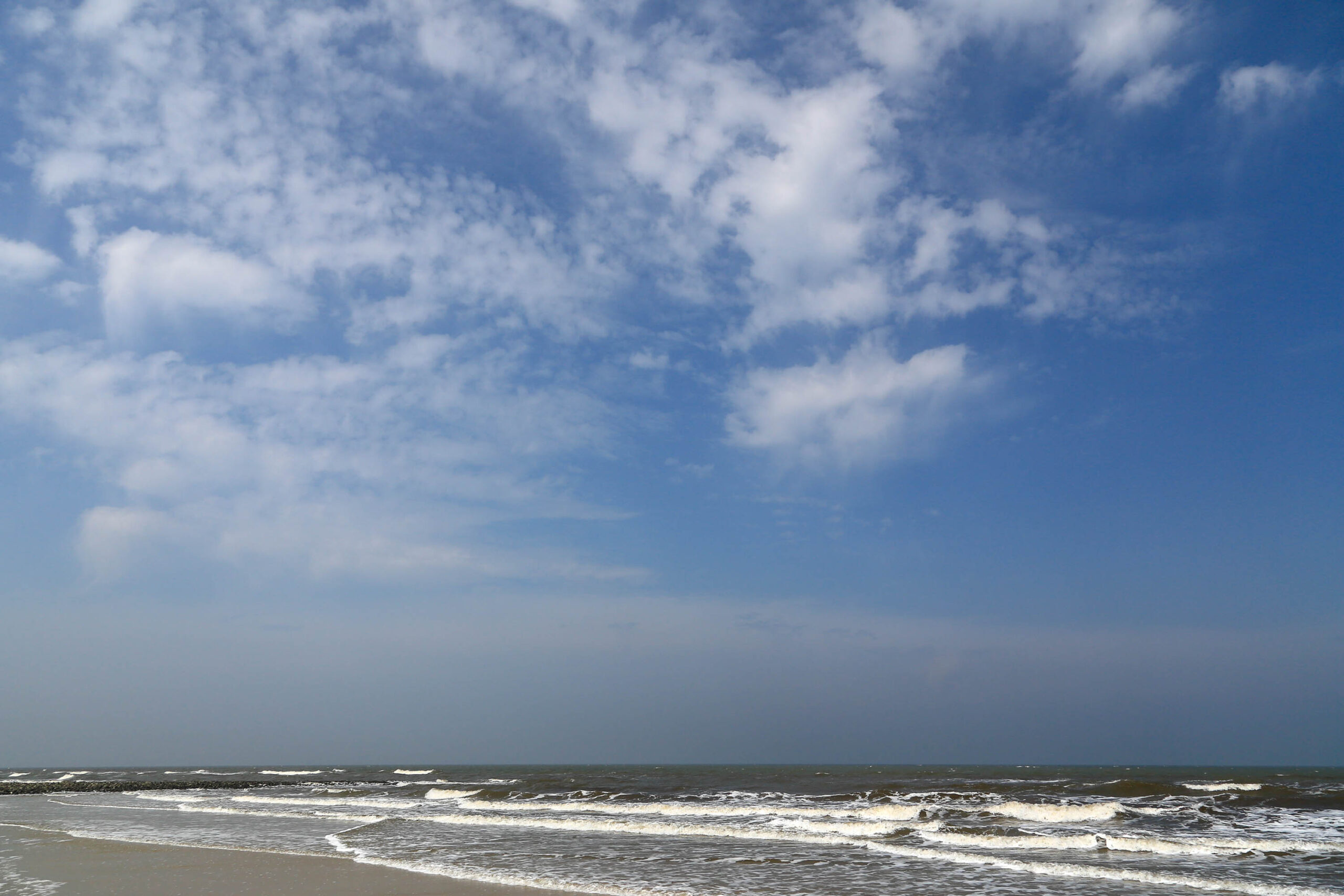 Die Aufnahme zeigt eine typische Szene an der Nordseeküste. Der Himmel ist tiefblau und von vereinzelten, weißen Cumuluswolken durchzogen. Das Meer ist aufgewühlt, mit brechenden Wellen, die am sandigen Strand zuschlagen. Der Strand selbst ist breit und sandig, mit einer leichten Neigung zum Wasser. Die Szene vermittelt ein Gefühl von Weite und Freiheit, typisch für die Küstenlandschaft der Nordsee.