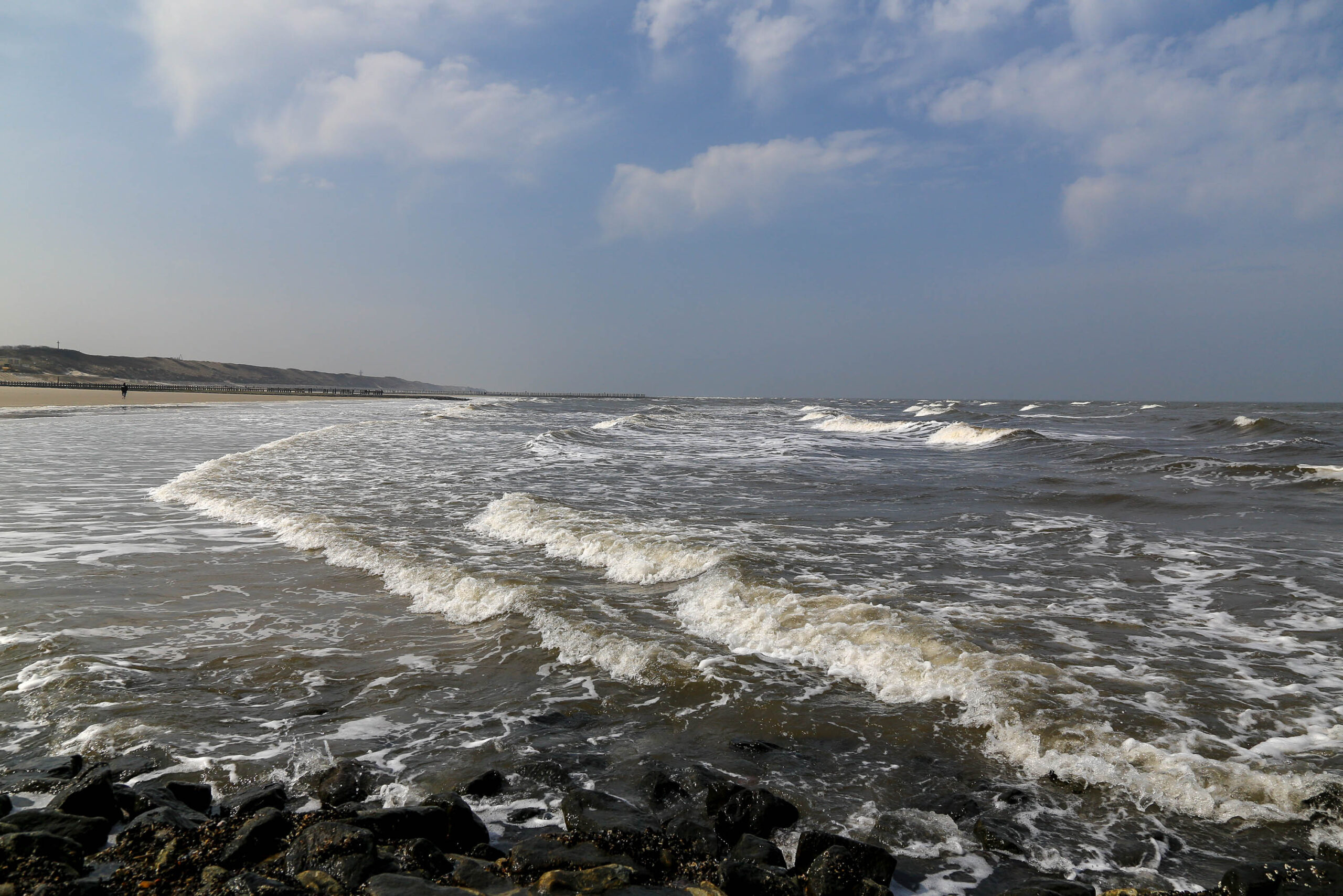 Die Aufnahme zeigt eine Szene vom Strand von Wangerooge, einer Insel in der Deutschen Bucht. Das Bild ist von einem Blickwinkel aus aufgenommen, der den weiten Blick über das Meer und den Strand betont. Das Wasser ist dunkel und aufgewühlt, mit weißen Schaumkronen, die auf die Küste treffen. Der Strand selbst ist aus dunklem Sand aufgebaut, der von Felsen unterbrochen wird. Im Hintergrund ist ein Deich zu erkennen, der die Insel vor dem Meer schützt. Einige Gebäude sind ebenfalls im Hintergrund sichtbar, die wahrscheinlich zu der kleinen Inselgemeinde gehören. Der Himmel ist bedeckt, aber es gibt Lichtdurchlässigkeit, die die Szene erhellt. Die Gesamtstimmung ist ruhig und friedlich, aber auch kraftvoll und dynamisch aufgrund der Wellenbewegung.