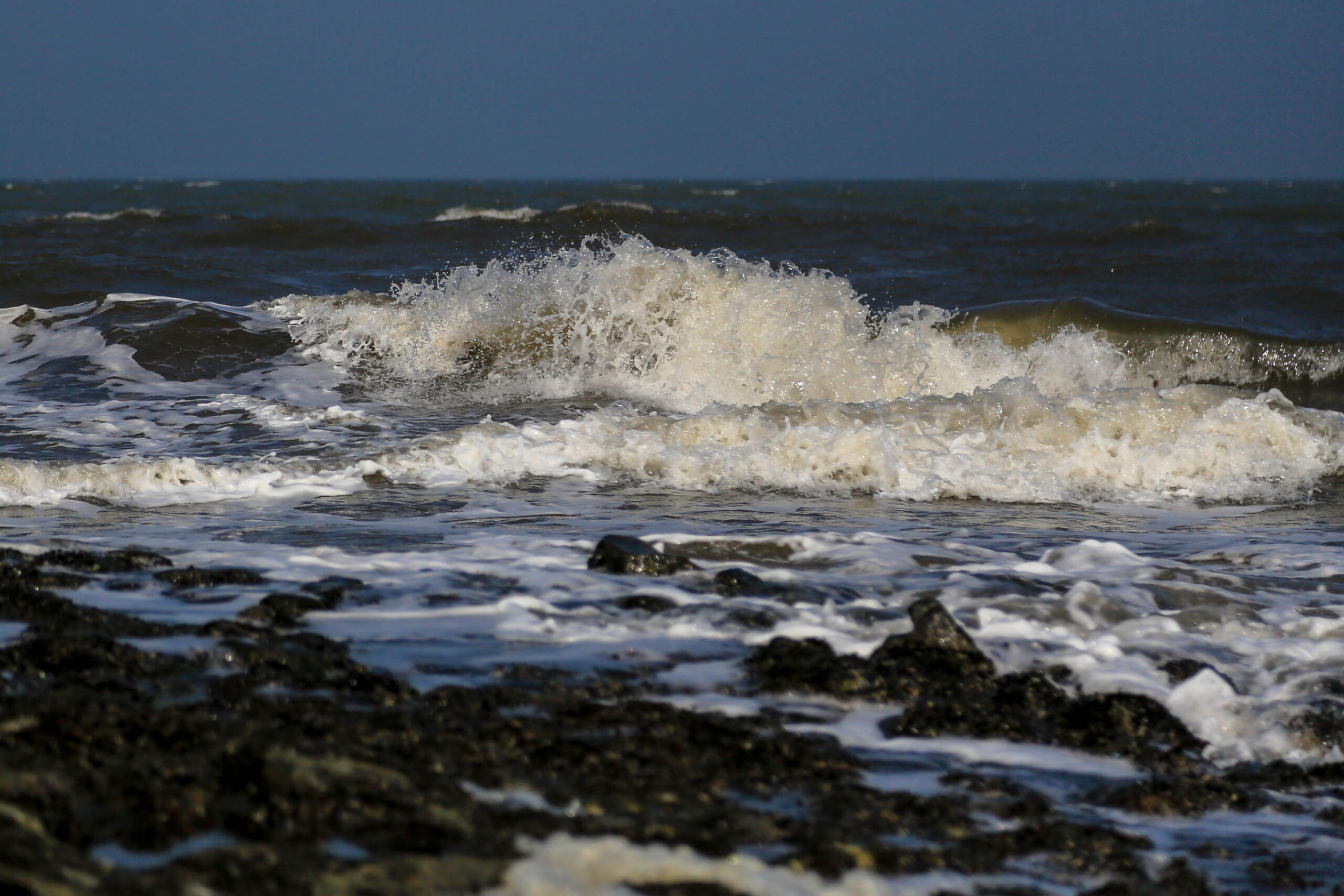 Das Bild zeigt eine dynamische Aufnahme von Wellen, die mit Kraft an einen felsigen Küstenstreifen schlagen. Das Wasser ist dunkelblau und tief, wobei die Wellenberge in hellem Weiß schäumen und brechen. Die Felsen sind dunkel und rau, was einen starken Kontrast zum schäumenden Wasser bildet. Die Aufnahme fängt die Energie und Kraft des Meeres ein. Die Szene vermittelt ein Gefühl von Bewegung und Dramatik. Die Aufnahme wurde im April 2018 auf der Insel Wangerooge in der Nordsee aufgenommen.