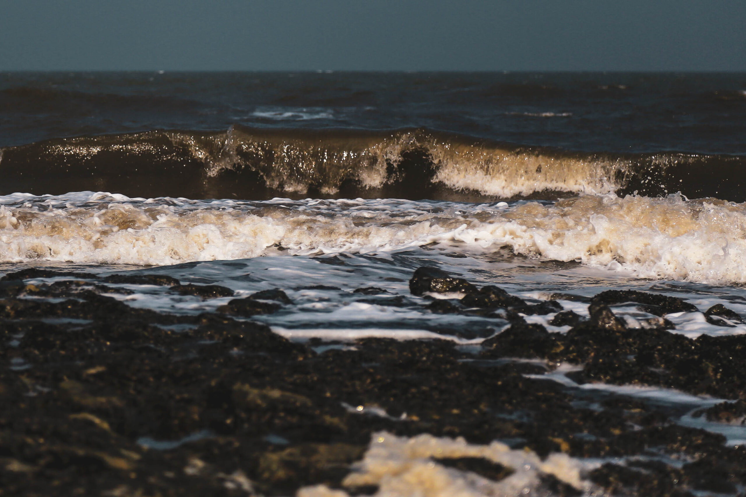 Das Bild zeigt eine dynamische Aufnahme von Wellen, die auf einen dunklen, felsigen Strand treffen. Das Wasser ist trüb und schäumt auf, während es über die Steine rollt. Die Farben sind gedämpft, mit einem starken Kontrast zwischen dem dunklen Strand und dem hellen Schaum des Wassers. Die Aufnahme fängt die Bewegung und Kraft des Meeres ein. Der Himmel ist dunkel und bewölkt, was die Szene dramatisch wirken lässt. Die Aufnahme wurde wahrscheinlich bei Ebbe aufgenommen, da der Strand weitläufig erscheint. Die Textur des Strandes ist deutlich sichtbar, mit vielen kleinen Steinen und Algen.