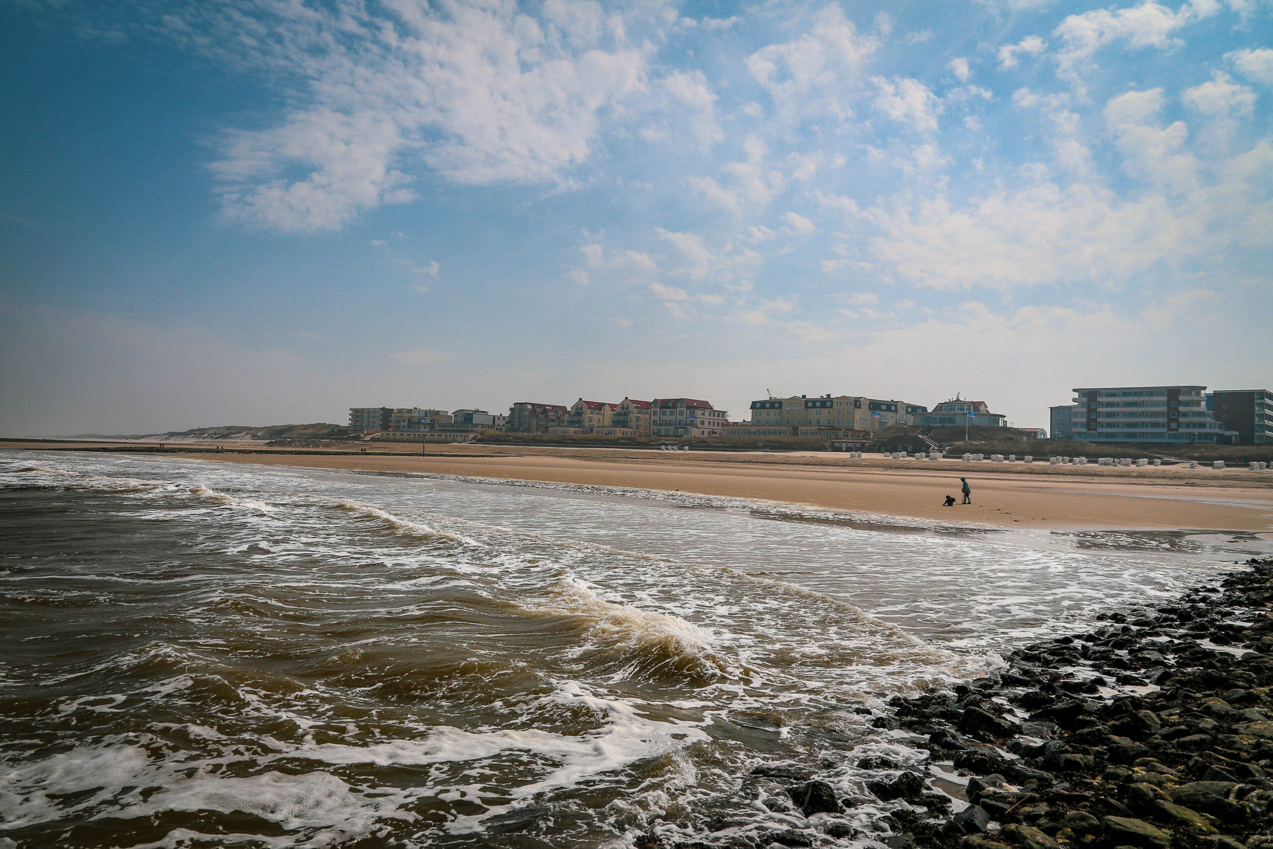 Das Foto zeigt einen weiten Blick auf einen Sandstrand an der Nordsee. Im Vordergrund brechen Wellen auf den Strand, wobei das Wasser eine bräunliche Farbe aufweist. Der Strand ist breit und sandig. Im Hintergrund sind mehrere Gebäude zu sehen, die typisch für einen Badeort sind. Der Himmel ist bewölkt, aber es gibt auch Lichtungen, die auf eine wechselhafte Wetterlage hindeuten. Eine einzelne Person ist im Hintergrund zu sehen, die am Strand spaziert. Die Szene vermittelt eine ruhige und friedliche Atmosphäre.