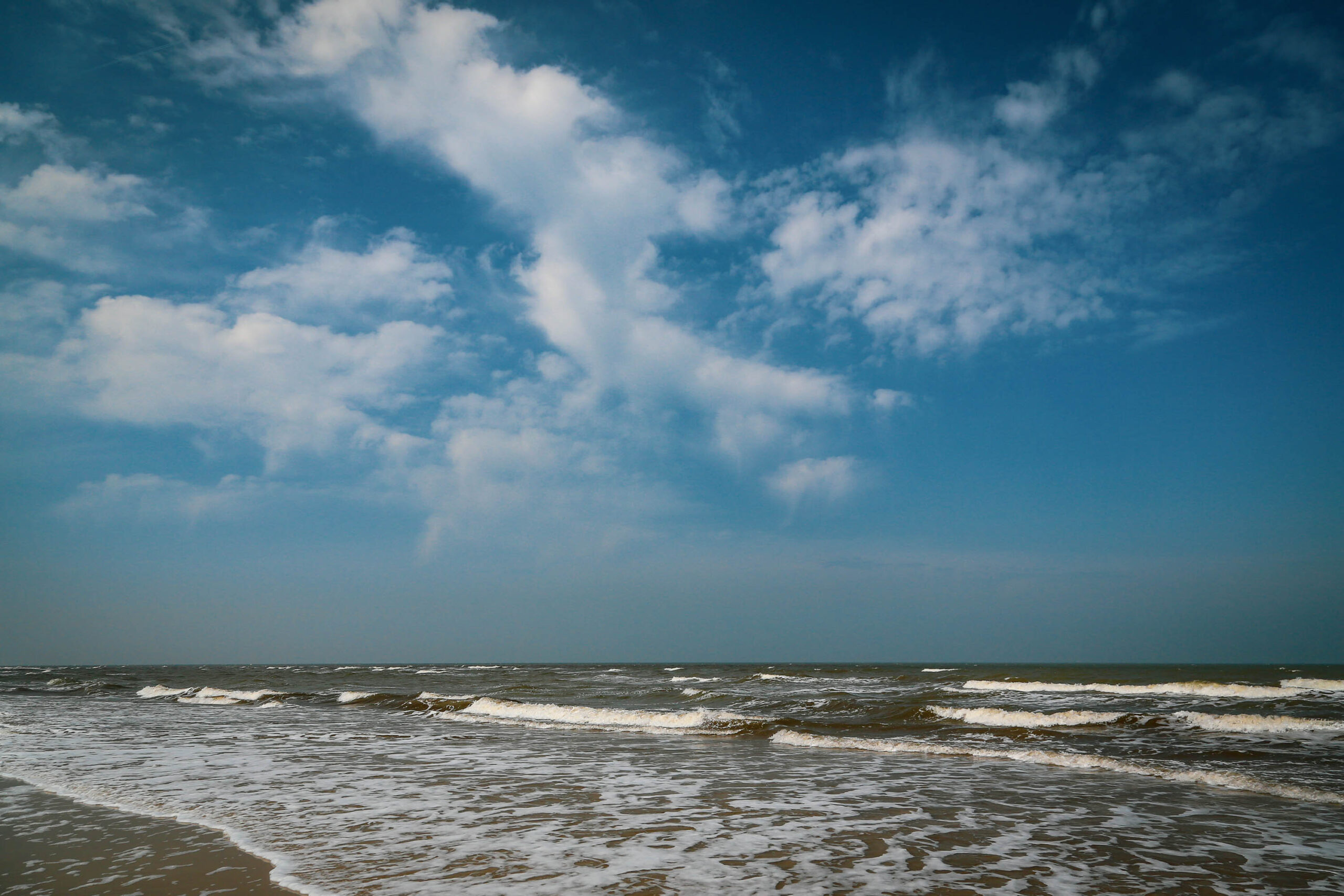 Die Aufnahme zeigt eine Weitwinkelaufnahme des Meeres an einem bewölkten Tag. Das Wasser ist dunkel und schäumt an der Küste. Der Himmel ist blau und von weißen Wolken bedeckt. Die Aufnahme wurde im April 2018 auf der Insel Wangerooge in der Nordsee aufgenommen.