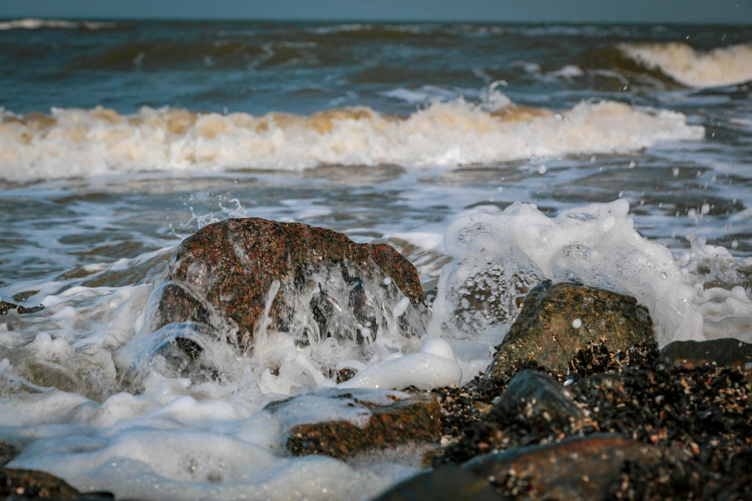 Die Aufnahme zeigt eine Nahaufnahme von Wellen, die über dunkle, moosbewachsene Felsen an einem Strand schlagen. Das Wasser ist trüb und schäumt, während es über die Felsen fließt. Im Hintergrund erstreckt sich das trübe, graue Meer. Die Szene vermittelt ein Gefühl von Bewegung und Kraft, während die Wellen unaufhörlich auf die Küste treffen. Die Farben sind gedämpft, mit einem Schwerpunkt auf Grau-, Braun- und Weißtönen. Die Aufnahme ist scharf und detailliert, wobei die Textur der Felsen und die Bewegung des Wassers gut zur Geltung kommen.