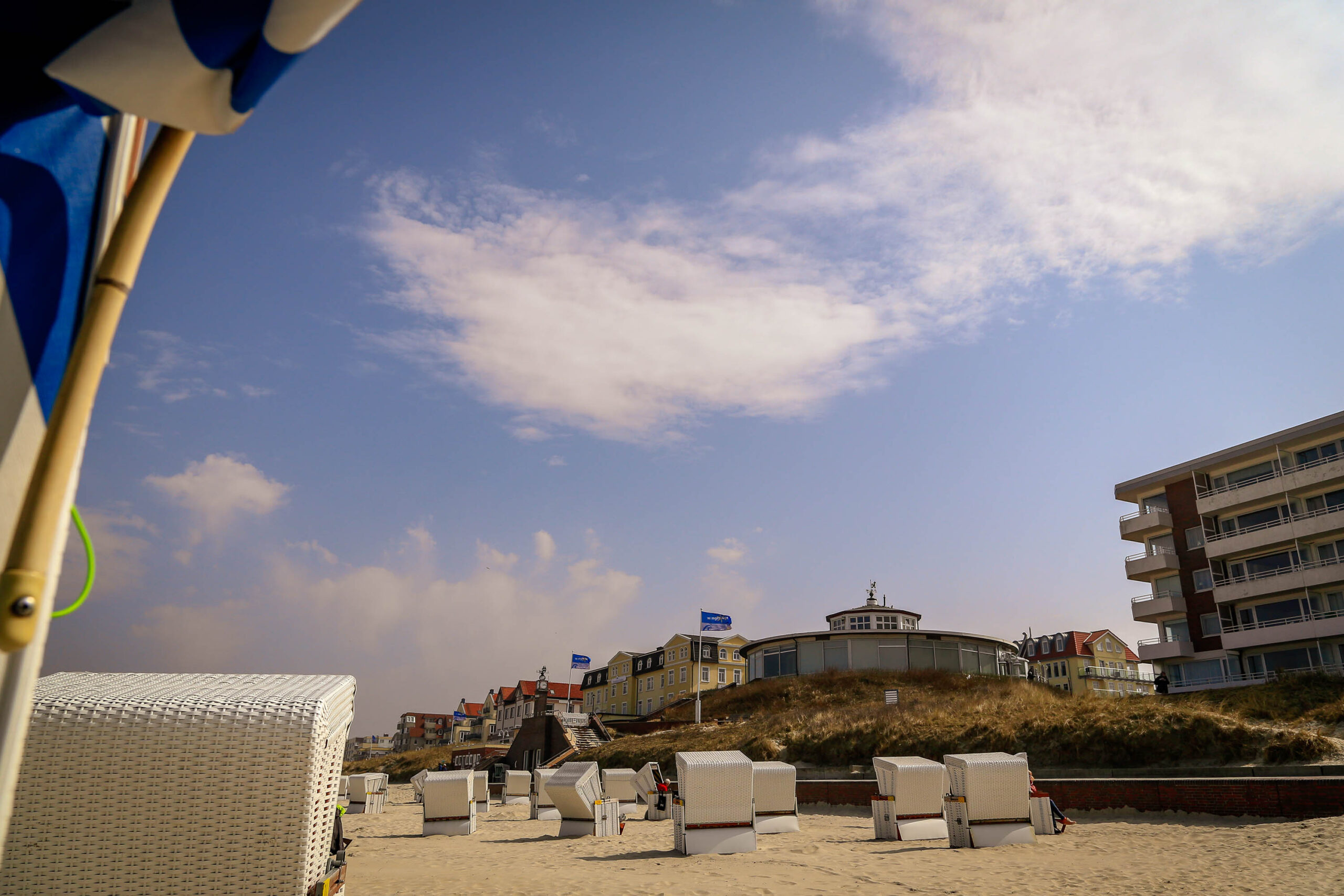 Die Aufnahme zeigt einen sonnigen Tag am Strand von Wangerooge. Im Vordergrund stehen zahlreiche Strandkörbe aus Rattan, die in einer Reihe angeordnet sind. Der Strand ist hell und sandig. Im Hintergrund erheben sich Gebäude, darunter ein markantes, rundes Gebäude mit einer Kuppel, sowie weitere Häuser und ein höher gelegener Gebäuderiegel. Der Himmel ist blau mit vereinzelten weißen Wolken. Die Perspektive ist von unten, was die Strandkörbe und die Gebäude betont. Die Aufnahme vermittelt eine entspannte und sonnige Atmosphäre.