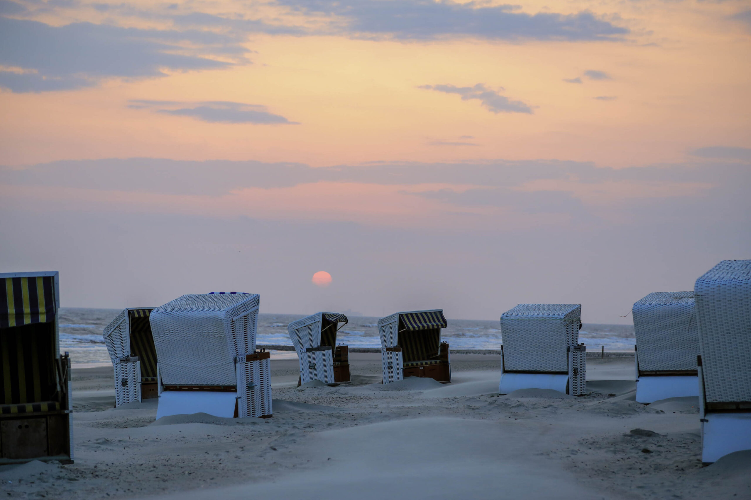 Das Bild zeigt eine Reihe von traditionellen Strandkörben auf dem Sandstrand der Insel Wangerooge. Die Körbe sind aus Rattan geflochten und stehen dicht aneinander. Der Himmel ist in warmen Farben von Rosa und Orange gehalten, was auf einen Sonnenaufgang oder Sonnenuntergang hindeutet. Im Hintergrund sind Wellen des Wattenmeeres zu sehen. Die Szene vermittelt eine ruhige und friedliche Atmosphäre.