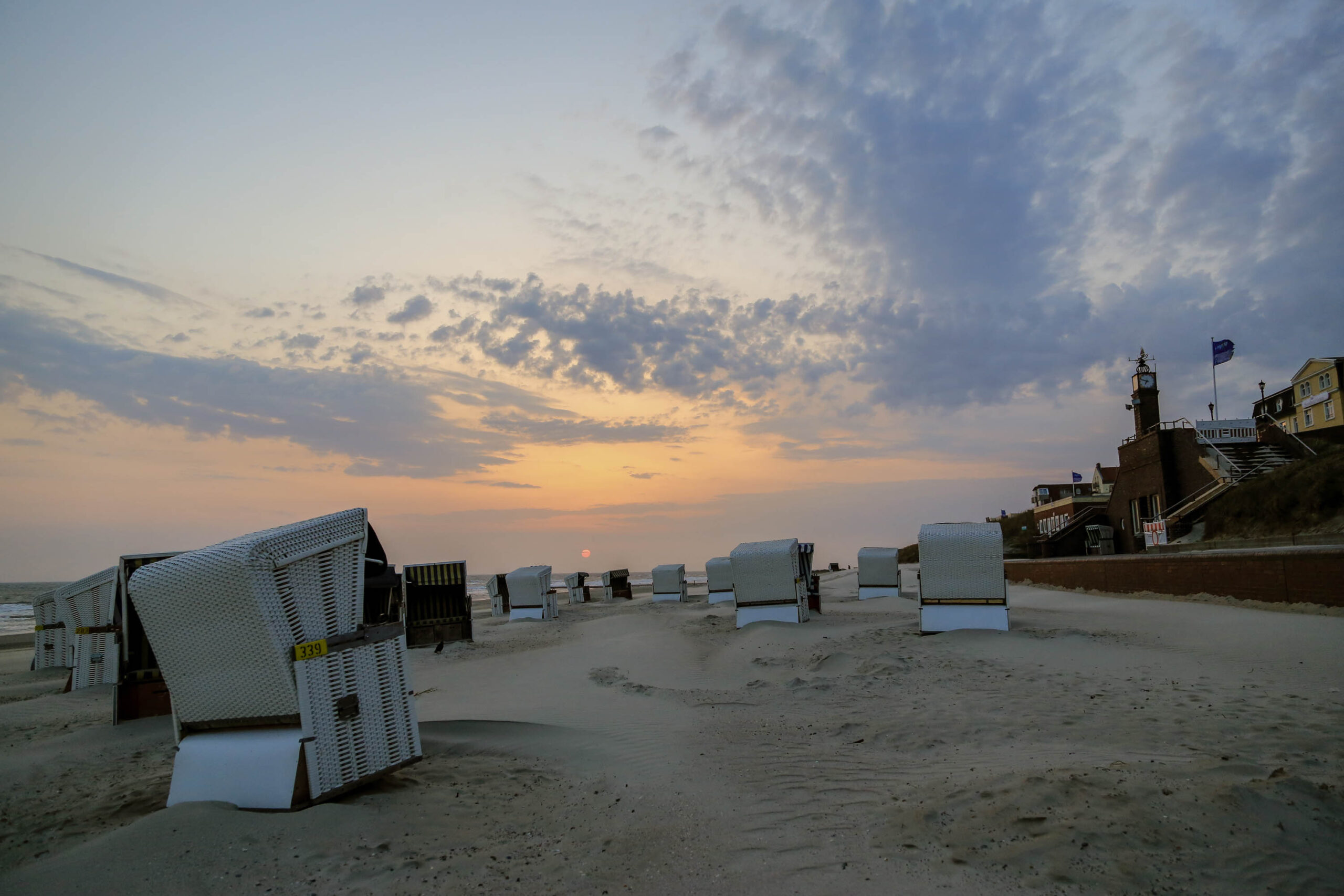 Das Bild zeigt eine friedliche Szene am Strand von Wangerooge bei Sonnenuntergang. Eine Reihe von weißen Strandkörben steht ordentlich auf dem Sand. Im Hintergrund sind Gebäude und ein Leuchtturm zu erkennen, die die typische Architektur der Insel widerspiegeln. Der Himmel ist von einem warmen Farbenspiel aus Orange, Rosa und Violett durchzogen, was eine ruhige und entspannende Atmosphäre schafft. Der Sand ist hell und reflektiert das Licht des Sonnenuntergangs. Die gesamte Szene vermittelt ein Gefühl von Ruhe und Abgeschiedenheit.