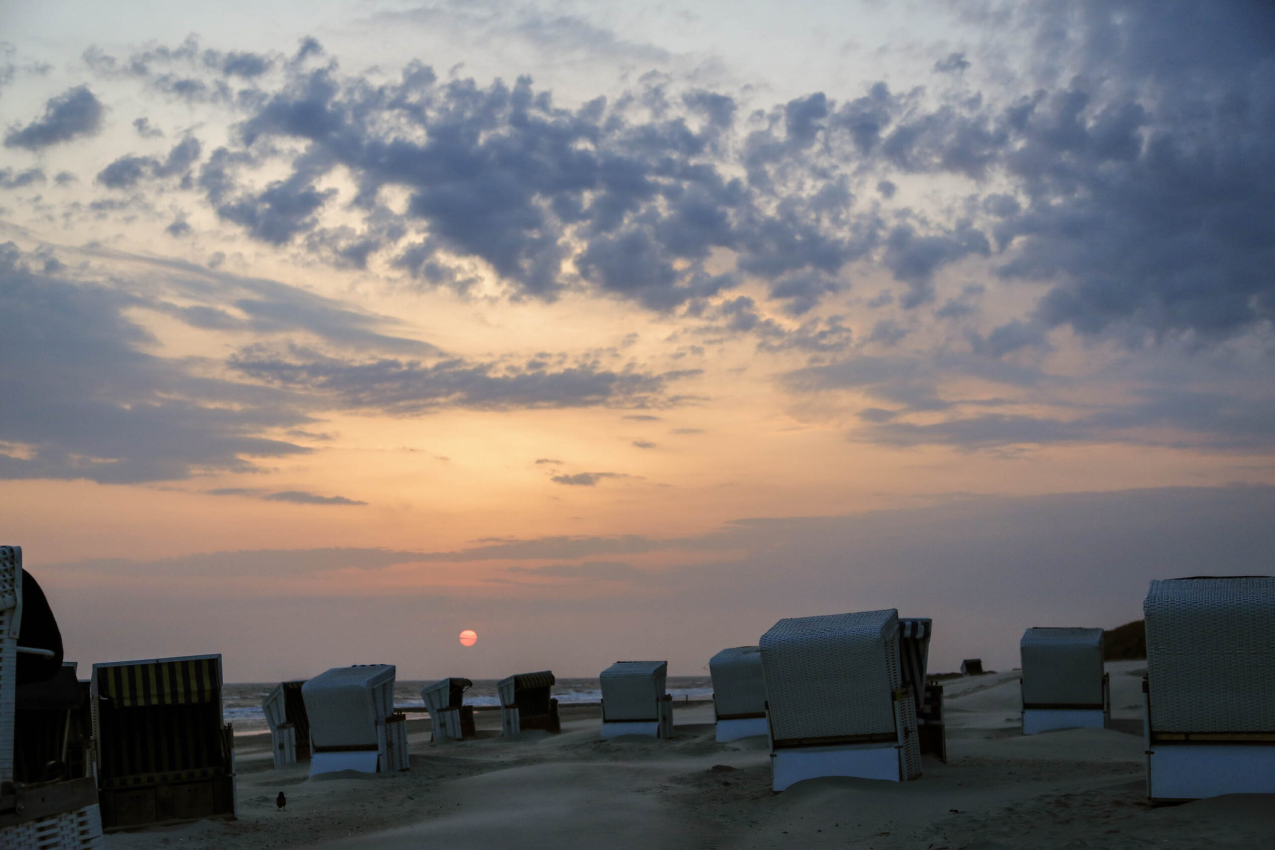 Das Bild zeigt einen Sonnenuntergang an einem Sandstrand auf der Insel Wangerooge. Im Vordergrund stehen mehrere traditionelle Strandkörbe, die aus Weidwerk bestehen und mit Stoffbezügen versehen sind. Der Sandstrand ist weitläufig und erstreckt sich bis zum Horizont. Der Himmel ist in warmen Orange- und Lilatönen gehalten, wobei die untergehende Sonne einen goldenen Schein erzeugt. Die Wolkendecke ist dynamisch und erzeugt ein dramatisches Lichtspiel. Die Szene vermittelt eine friedliche und entspannte Atmosphäre.
