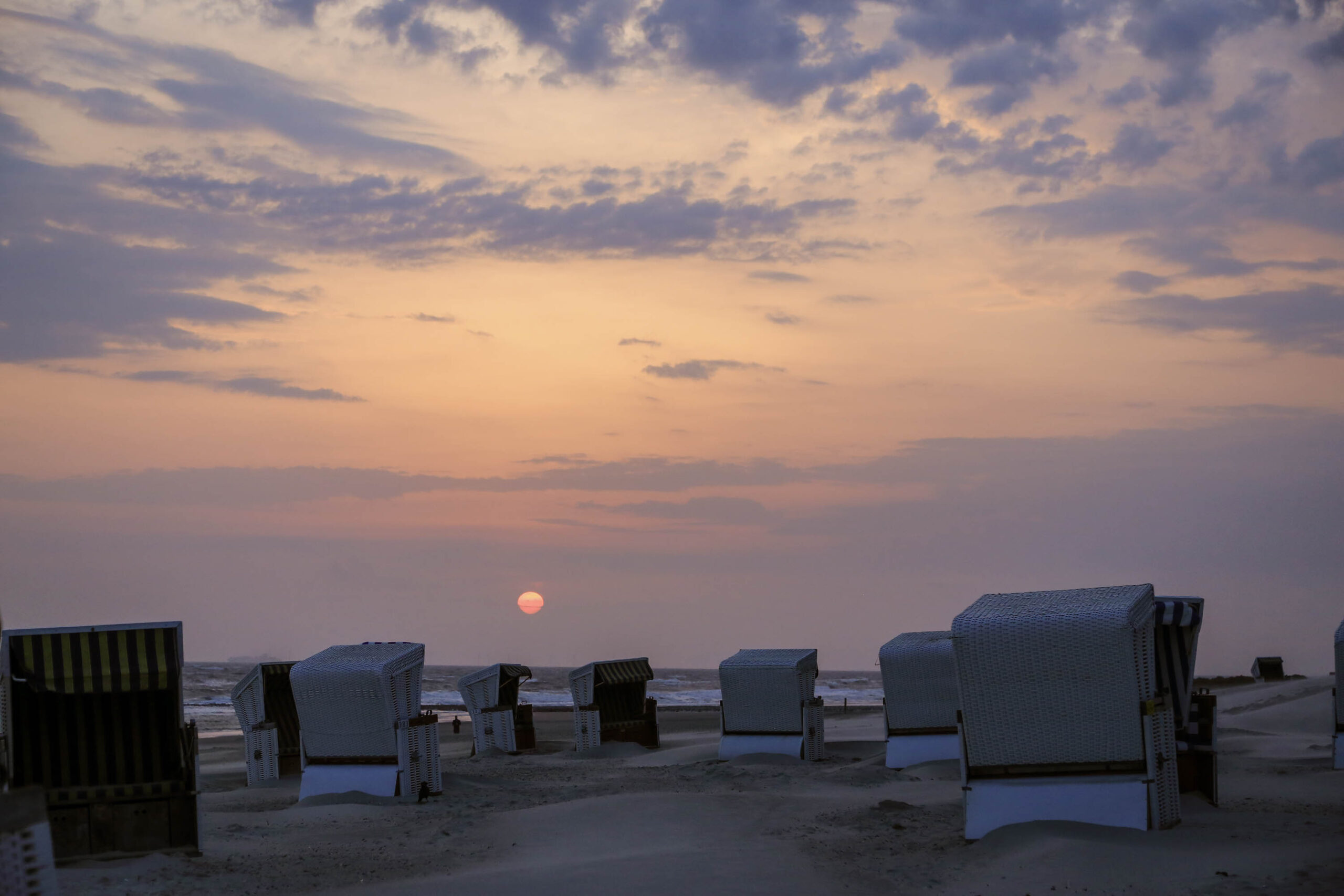 Das Bild zeigt eine Reihe von traditionellen Strandkörben, die auf einem Sandstrand stehen. Der Himmel ist in warmen Orange- und Lilatönen gehalten, was auf einen Sonnenuntergang hindeutet. Die Strandkörbe sind aus Rattan geflochten und wirken in der Abendsonne sehr idyllisch. Der Strand ist weitläufig und leer, was eine ruhige und friedliche Atmosphäre schafft. Die Szene vermittelt ein Gefühl von Ruhe und Entspannung.