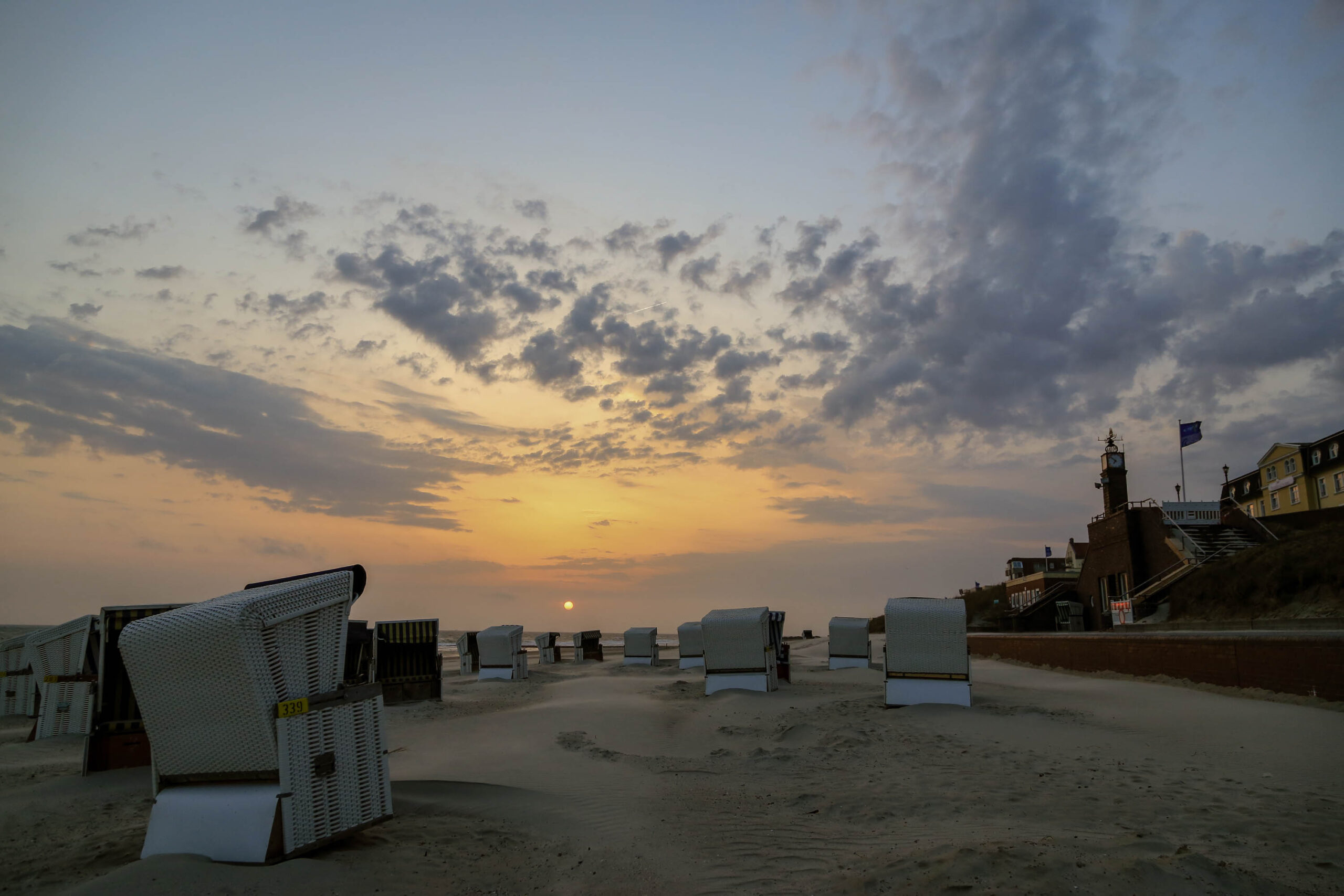 Das Bild zeigt einen Sandstrand bei Sonnenuntergang. Zahlreiche Strandkörbe sind in einer Reihe aufgestellt und leiten den Blick in die Ferne. Im Hintergrund sind Gebäude zu erkennen, die vermutlich zu Wangerooge gehören. Der Himmel ist bedeckt mit Wolken, die das Licht der untergehenden Sonne reflektieren und eine dramatische Atmosphäre erzeugen. Die Szene vermittelt ein Gefühl von Ruhe und Abgeschiedenheit.