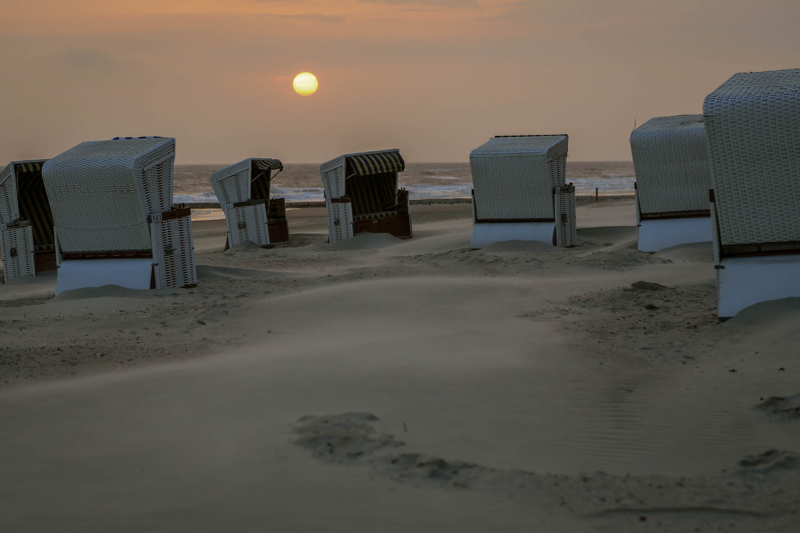 Das Foto zeigt eine Reihe von traditionellen Strandkörben auf der Insel Wangerooge, Nordsee. Die Körbe sind aus Weidwerk gefertigt und stehen dicht aneinander auf einem Sandstrand. Der Himmel ist in warmen Orange- und Gelbtönen gehalten, was auf einen Sonnenuntergang hindeutet. Der Sand ist fein und hell, und die Szene wirkt ruhig und friedlich. Die Körbe sind typisch für die Küstenregionen Deutschlands und bieten einen geschützten Platz zum Entspannen am Strand.