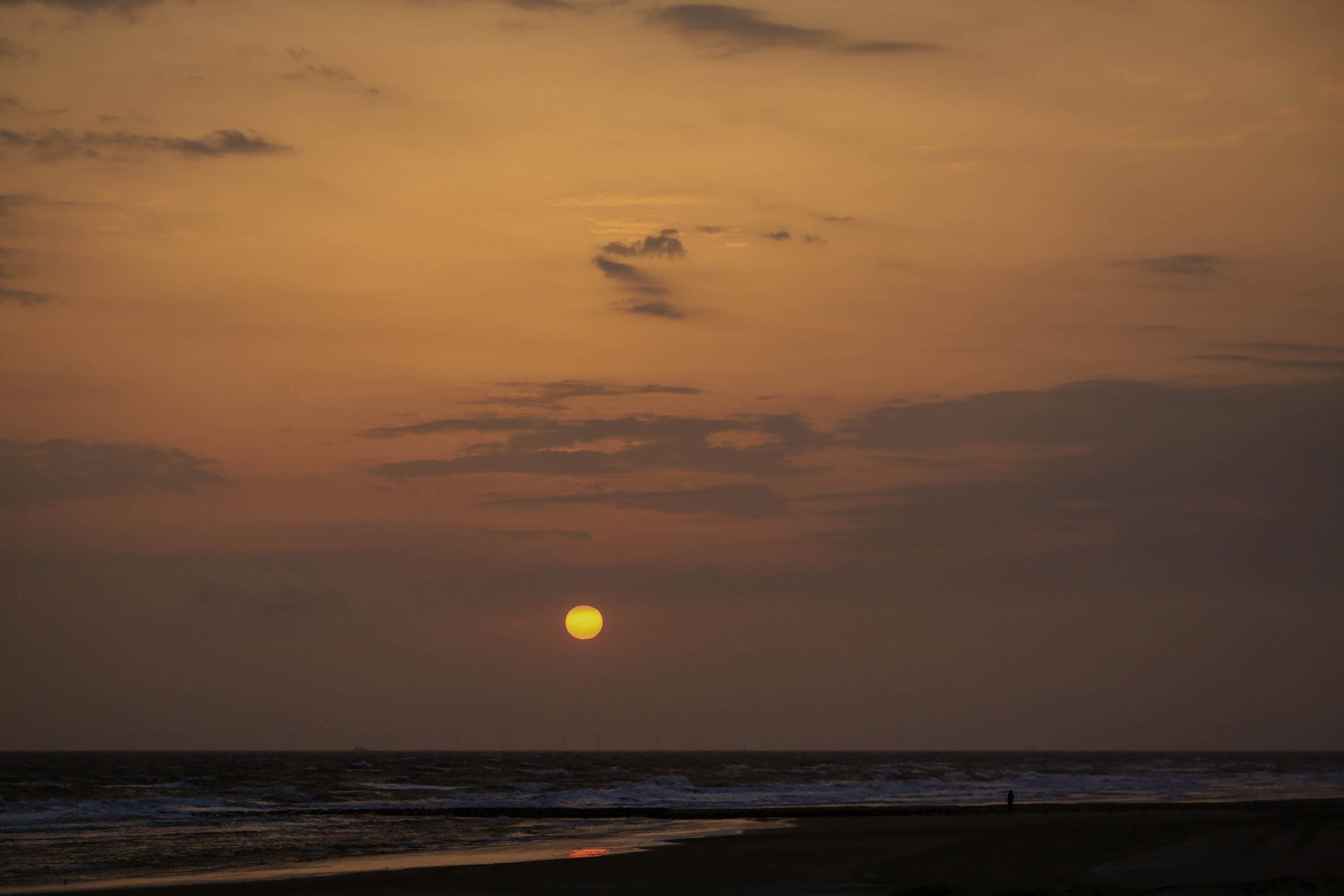 Das Bild zeigt einen Sonnenuntergang über dem Meer. Die Sonne ist als heller Kreis am Horizont zu sehen, umgeben von einem orangefarbenen Himmel. Das Meer ist ruhig und spiegelt das Licht der Sonne wider. Am Strand befindet sich eine einzelne Person, die in Richtung des Sonnenuntergangs geht. Der Himmel ist mit vereinzelten Wolken bedeckt, die das Licht der Sonne streuen.