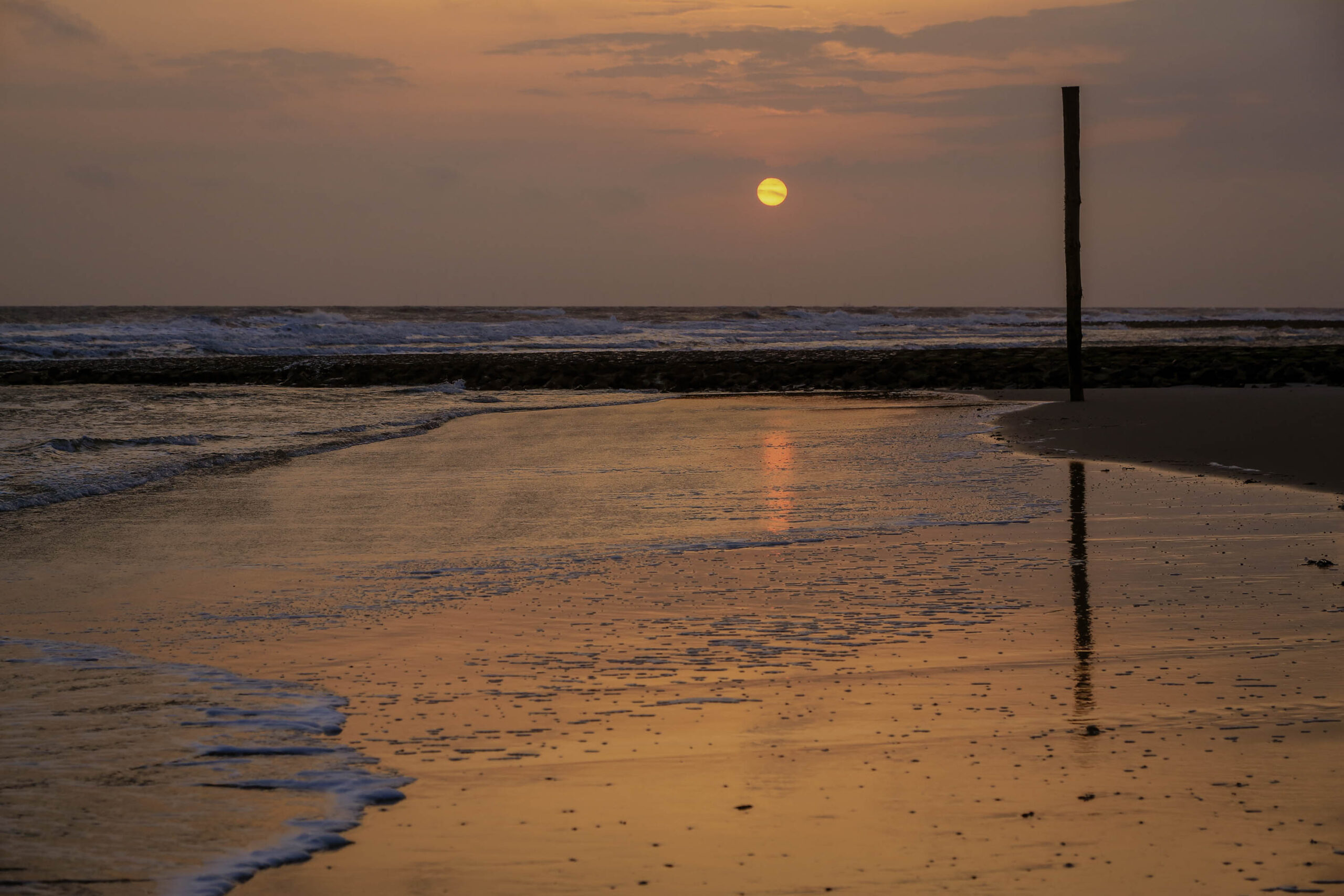 Das Bild zeigt einen Sonnenuntergang über dem Meer, aufgenommen auf der Insel Wangerooge. Der Himmel ist in warmen Orange- und Gelbtönen gehalten, die sich im feuchten Sand spiegeln. Im Vordergrund erstreckt sich eine breite Sandfläche, die von kleinen Wellen berührt wird. Ein einzelner, schlanker Pfosten steht vertikal im Sand und spiegelt sich ebenfalls im Wasser. Die Szene vermittelt eine ruhige und friedliche Atmosphäre.