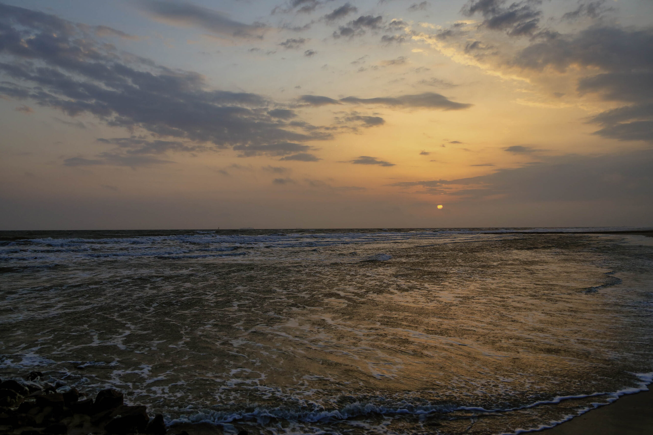 Das Bild zeigt einen Sonnenuntergang über dem Meer, aufgenommen auf der Insel Wangerooge. Der Himmel ist in warmen Gelb- und Orangetönen gehalten, während das Wasser in dunkleren Brauntönen schimmert. Die Sonne steht tief am Horizont und erzeugt einen goldenen Schein auf der Wasseroberfläche. Die Wellen brechen sanft am Strand. Die Szene vermittelt eine friedliche und ruhige Atmosphäre.