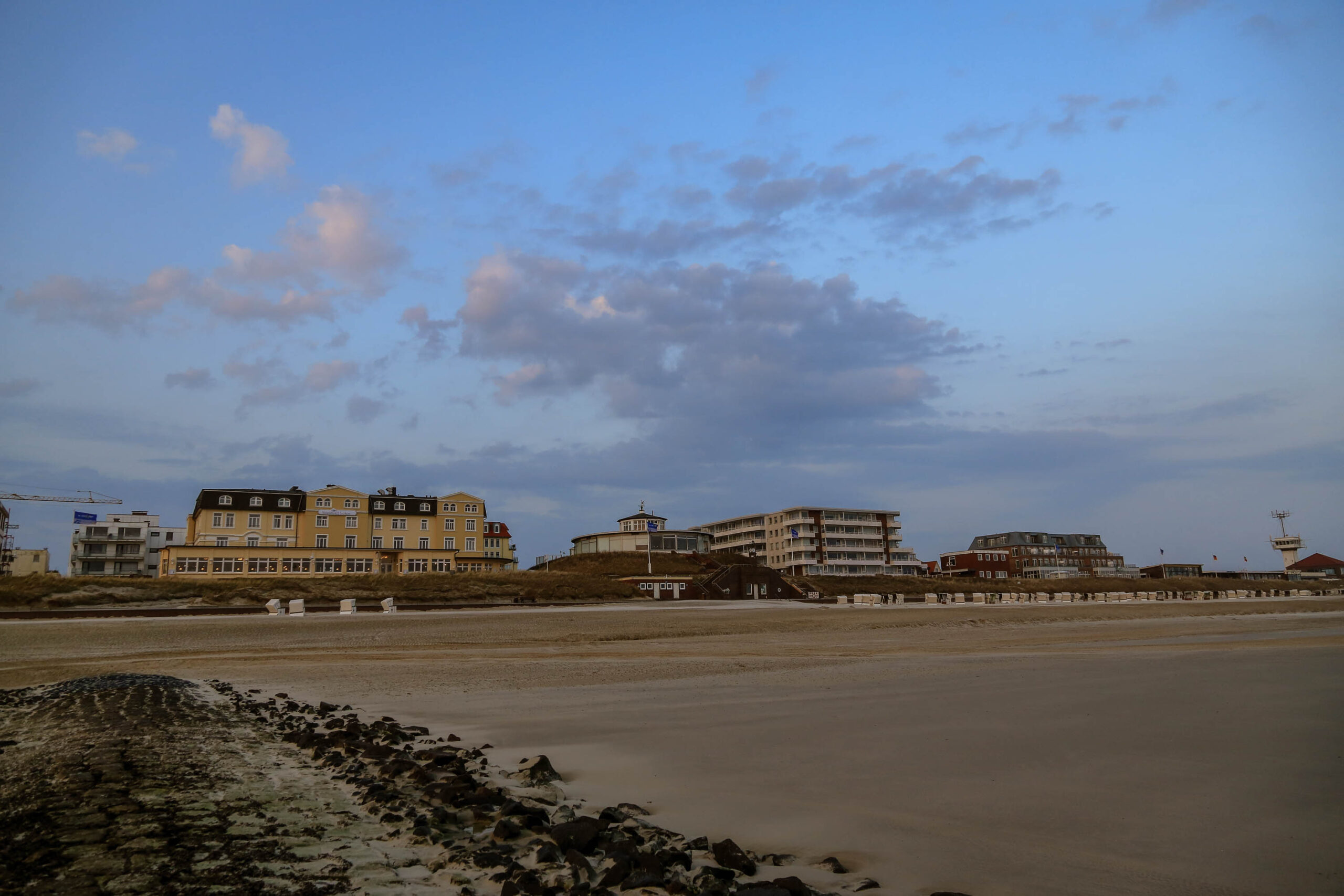Die Aufnahme zeigt eine typische Szene von Wangerooge, einer Insel in der Deutschen Nordsee. Die Gebäude im Hintergrund sind Hotelkomplexe und Ferienwohnungen, die für den Tourismus gebaut wurden. Der Sandstrand erstreckt sich weitläufig und wird von einigen Felsen unterbrochen. Der Himmel ist in warmen Farben gehalten, was auf einen Sonnenuntergang hindeutet. Die Aufnahme vermittelt eine ruhige und friedliche Atmosphäre.