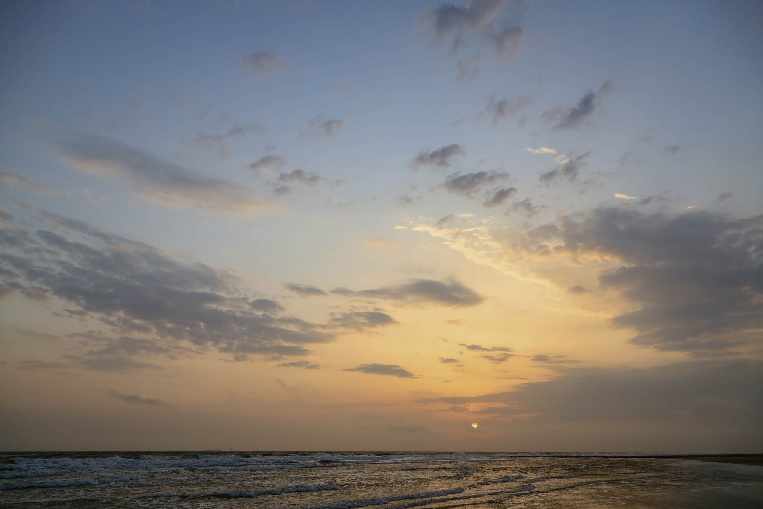 Das Bild zeigt einen Sonnenuntergang über dem Meer. Der Himmel ist in warme Farben wie Gelb und Orange getaucht, mit vereinzelten weißen Wolken. Der Horizont ist kaum erkennbar, da er durch den Dunst und die Wellen verschwimmt. Das Wasser ist dunkelblau und reflektiert das Licht des Himmels. Der Strand ist sandig und leicht feucht. Die Szene vermittelt eine ruhige und friedliche Atmosphäre.