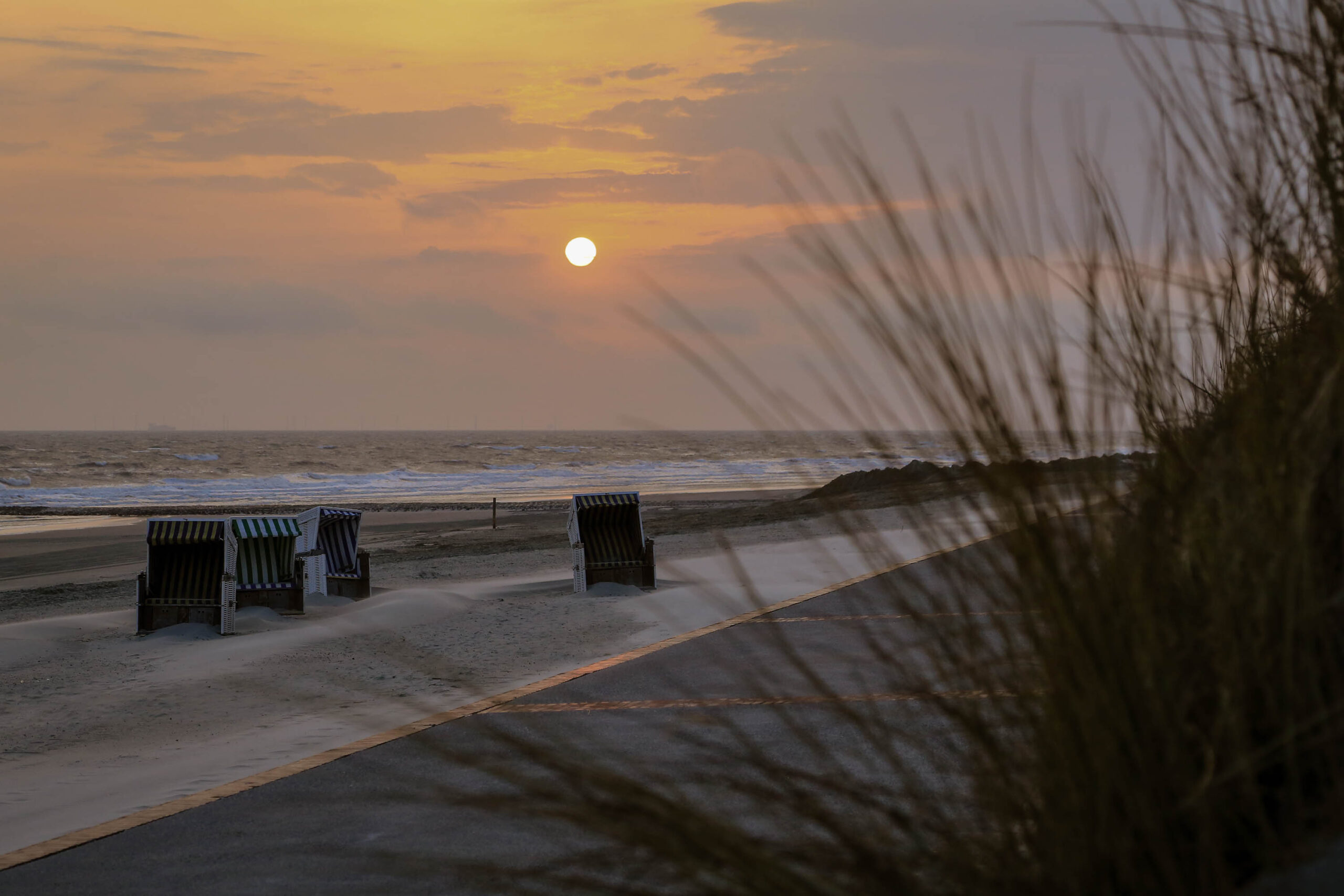 Das Bild zeigt einen Sonnenuntergang an einem Strand auf der Insel Wangerooge. Im Vordergrund stehen mehrere Strandkörbe, die sich in der warmen Abendsonne präsentieren. Die Dünen ragen im Hintergrund auf, und das Meer erstreckt sich bis zum Horizont. Der Himmel ist in warmen Orange- und Gelbtönen gehalten, was eine friedliche und entspannte Atmosphäre schafft. Die Silhouette von Gräsern im Vordergrund verleiht dem Bild eine zusätzliche Ebene der Tiefe und Textur.