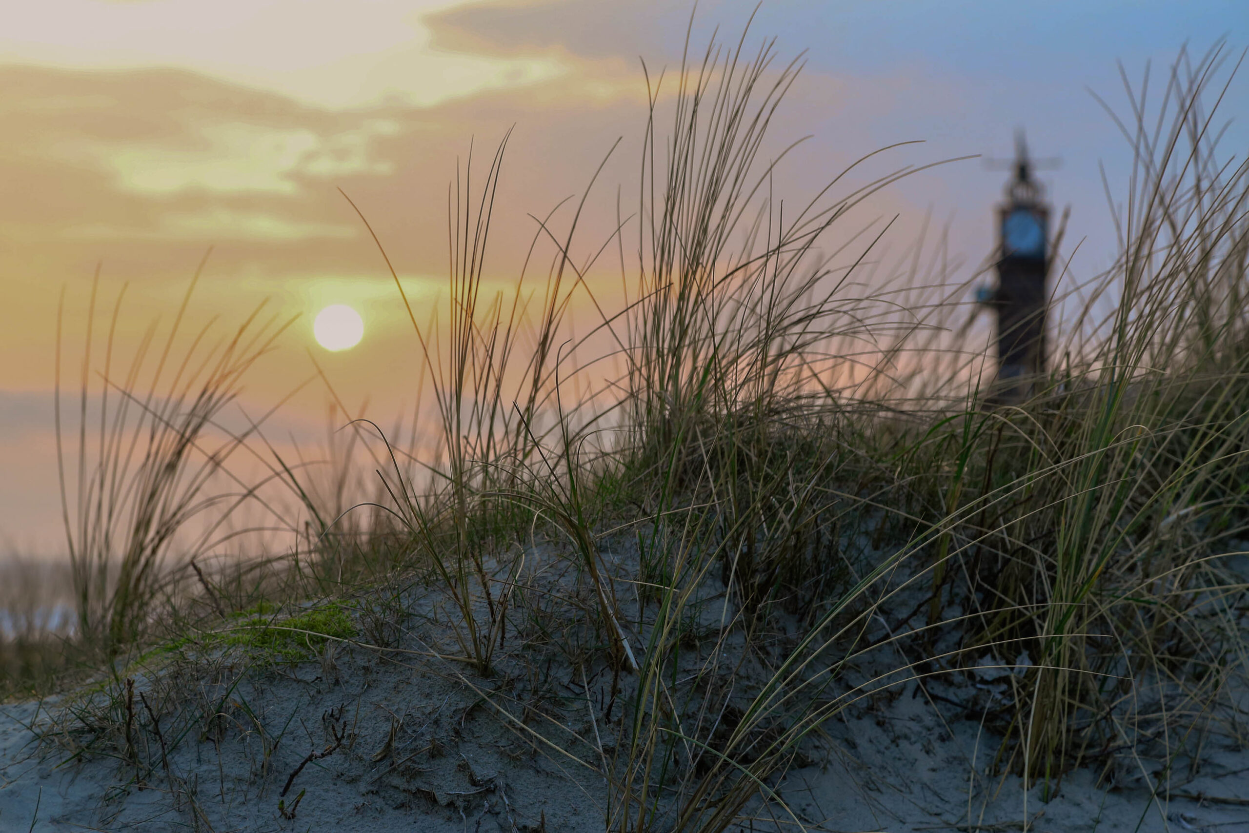 Das Bild zeigt einen Sonnenaufgang über der Insel Wangerooge in der Nordsee. Im Vordergrund erheben sich Dünen, die mit Grasbüschen bewachsen sind. Im Hintergrund ist der Leuchtturm von Wangerooge zu sehen, der leicht im Dunst liegt. Der Himmel ist in warmen Farben wie Orange, Rosa und Blau gemalt, was eine friedliche und ruhige Atmosphäre schafft. Das Bild fängt die Schönheit der norddeutschen Küstenlandschaft ein.