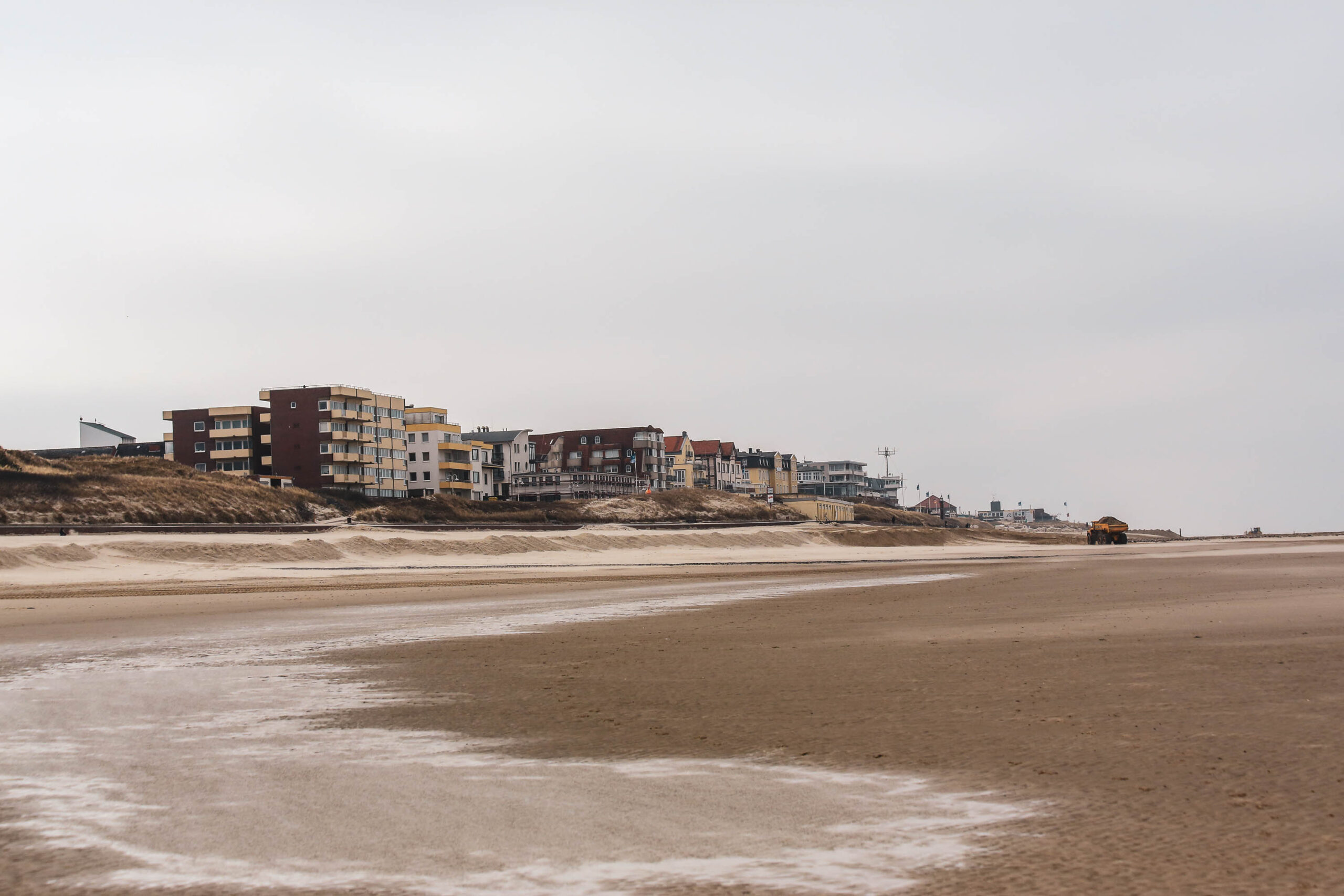 Die Aufnahme zeigt eine typische Küstenlandschaft auf der Insel Wangerooge in der Nordsee. Eine Reihe von Häusern, die in verschiedenen Farben und Stilen gehalten sind, erstreckt sich entlang des Sandstrandes. Die Häuser sind in unterschiedlichen Höhen angeordnet und scheinen in die Dünen eingebettet zu sein. Der Strand ist weitläufig und sandig, mit deutlichen Spuren von Gezeiten. Der Himmel ist bedeckt und erzeugt eine gedämpfte, ruhige Atmosphäre. Die Aufnahme fängt die charakteristische Schönheit und Ruhe einer norddeutschen Insel ein.