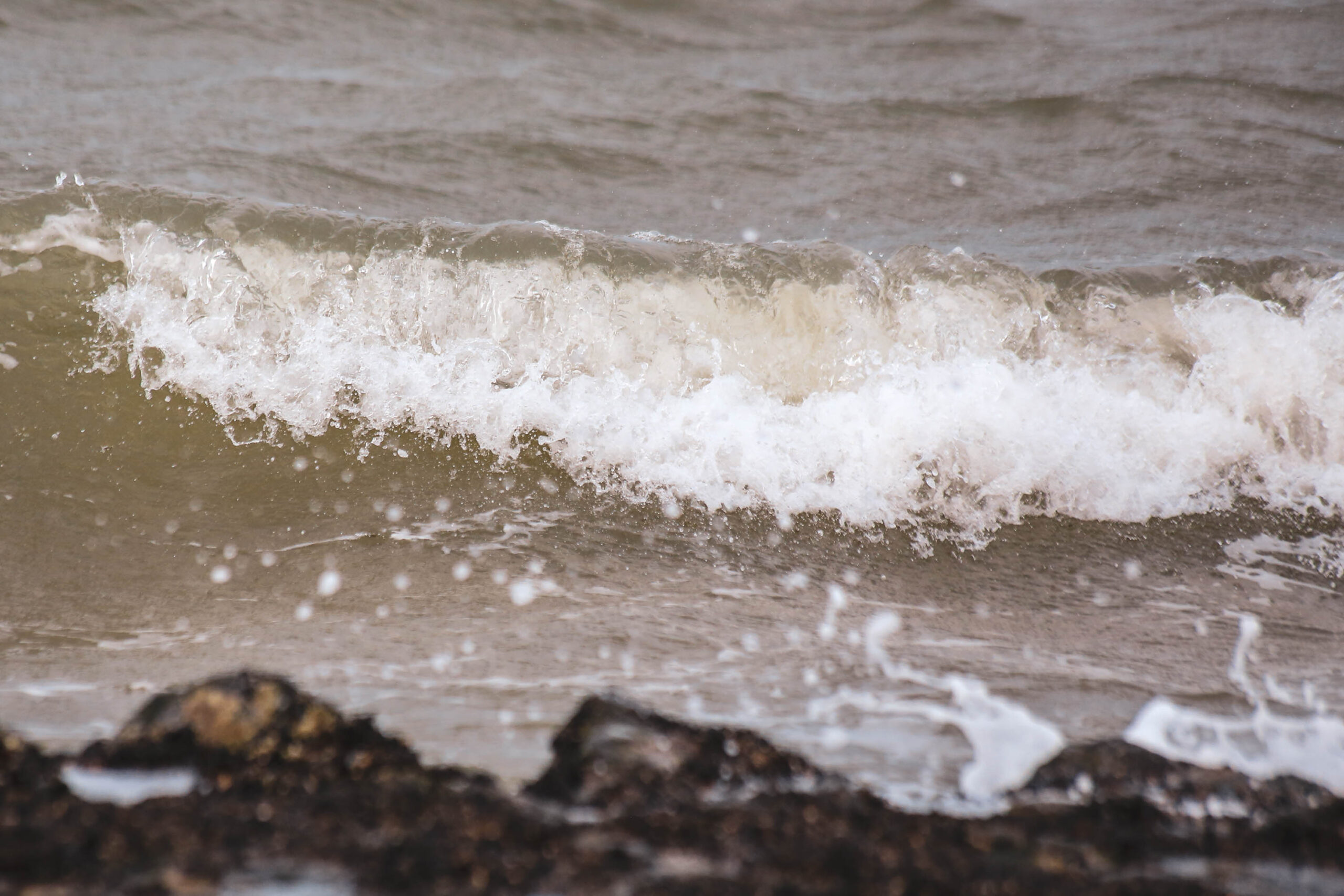 Das Bild zeigt eine Nahaufnahme einer Welle, die an einen dunklen, steinigen Strand trifft. Das Wasser ist trüb und braun, mit weißen Schaumkronen. Die Textur des Wassers ist deutlich sichtbar, mit kleinen Bläschen und Wirbeln. Der Strand besteht aus dunklen Steinen und Kies. Die Szene vermittelt eine Atmosphäre von Ruhe und Bewegung.