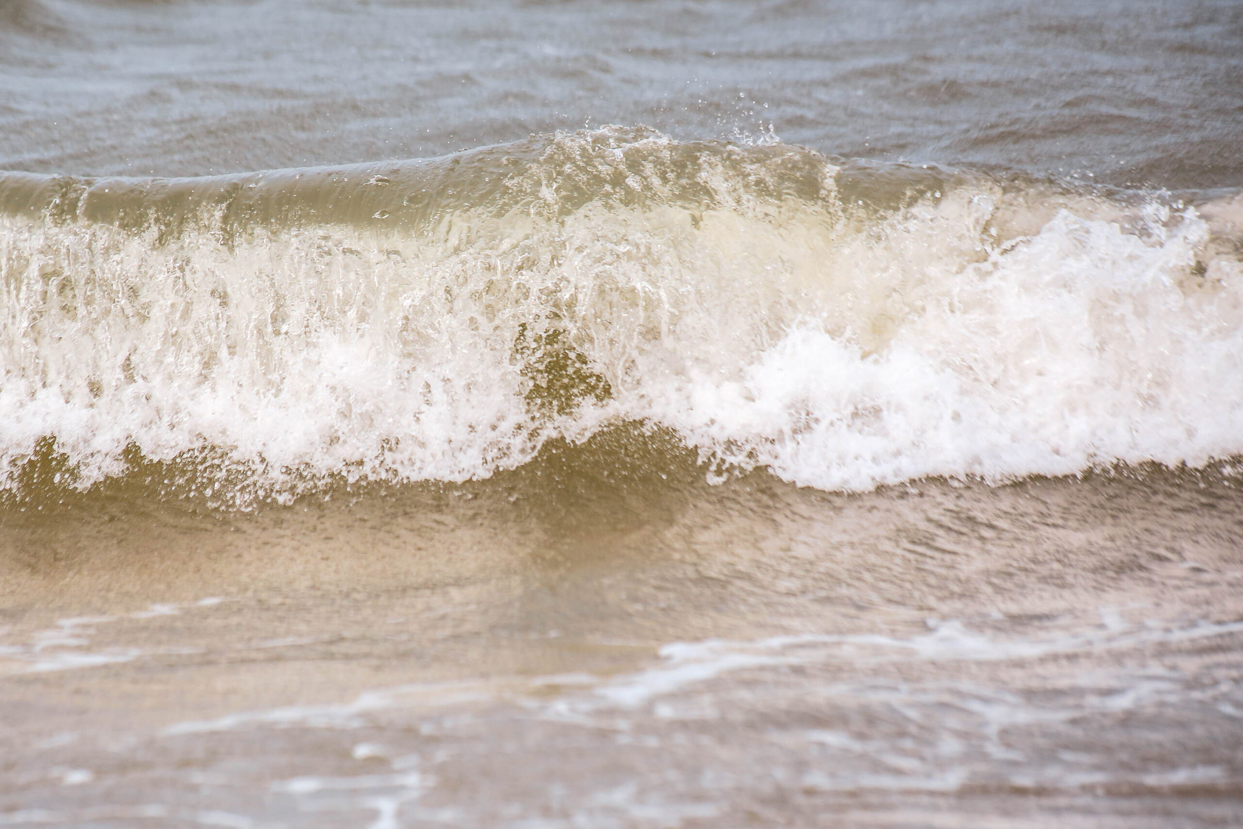 Das Bild zeigt eine Nahaufnahme einer Welle, die sich auf einen sandigen Strand rollt. Das Wasser ist trüb und sandig, mit weißen Schaumkronen an der Wellenkante. Der Sand unter dem Wasser ist ebenfalls sandig und trüb. Das Bild vermittelt ein Gefühl von Bewegung und Ruhe.