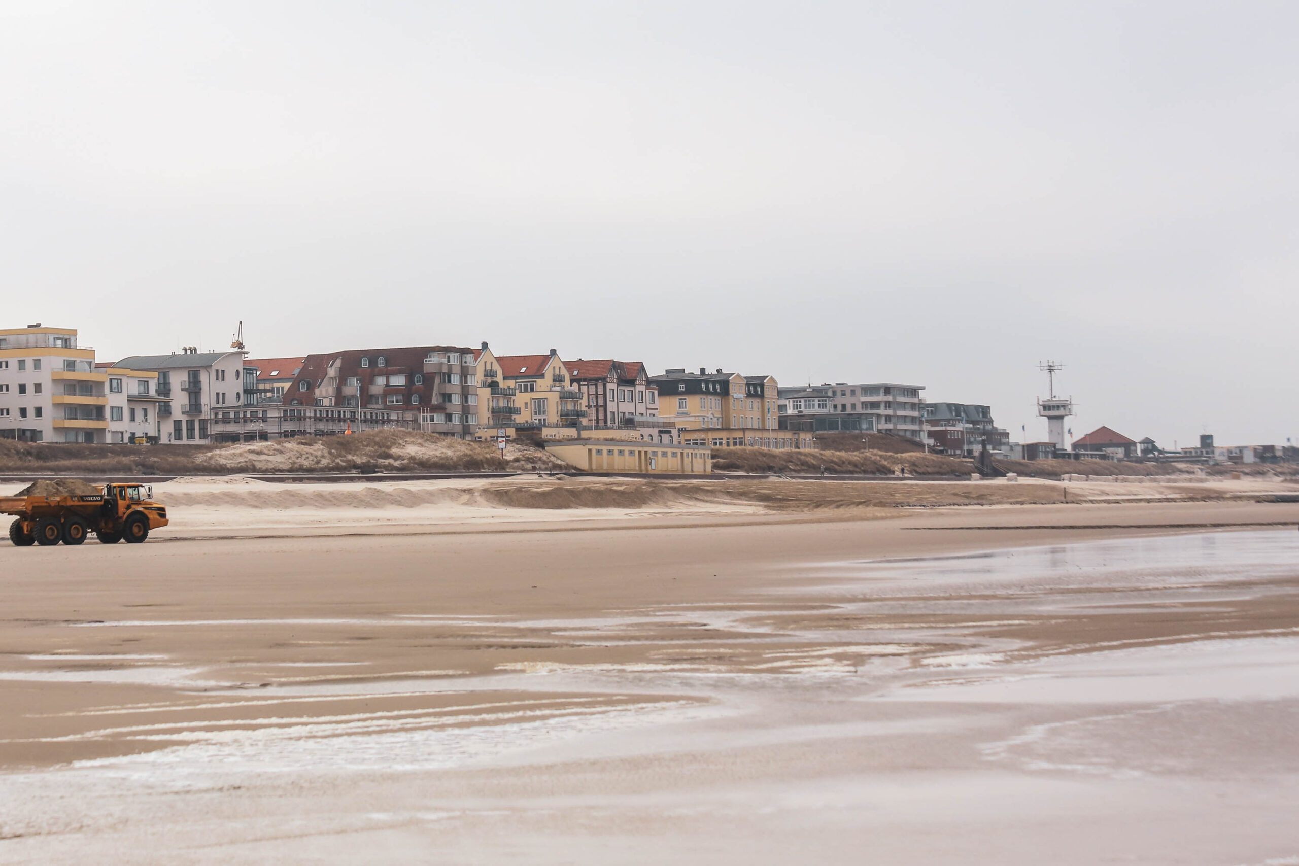 Die Aufnahme zeigt eine typische Szene von Wangerooge, einer Insel in der Nordsee. Ein breiter Sandstrand erstreckt sich bis zum Horizont, bedeckt von einer dünnen Schicht aus feuchtem Sand. Ein gelber Bagger fährt auf dem Strand. Im Hintergrund reihen sich Häuser aneinander, die typisch für die Architektur von Inseln in der Nordsee sind. Ein Leuchtturm ragt in den Himmel. Der Himmel ist bedeckt und die Lichtverhältnisse sind diffus.