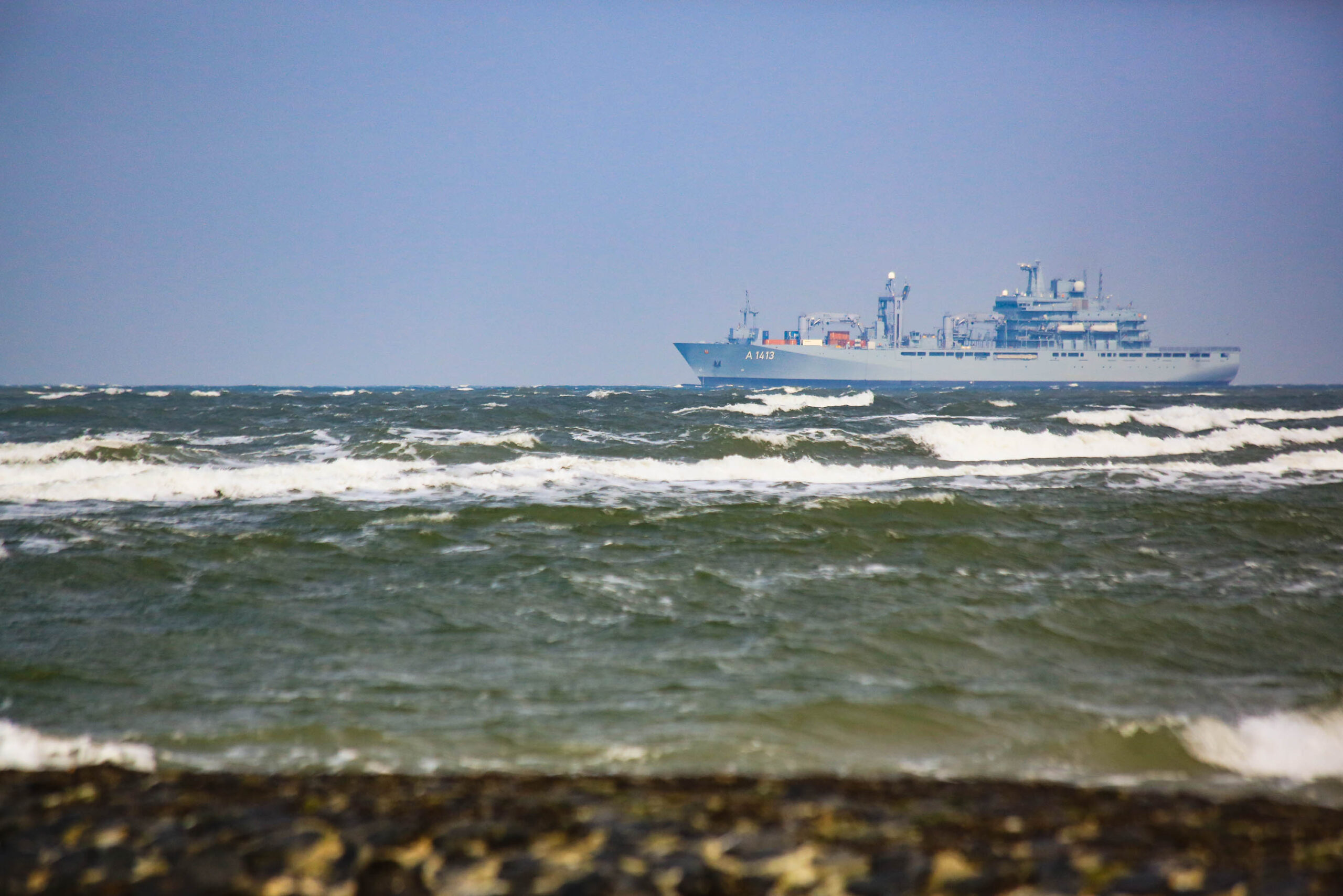 Das Bild zeigt ein großes, graues Kriegsschiff, das sich durch stürmische See bewegt. Die Wellen sind hoch und brechen, was die Szene dynamisch und kraftvoll wirken lässt. Der Himmel ist bedeckt und der Horizont ist kaum erkennbar. Im Vordergrund sind dunkle Felsen oder Steine zu sehen, die den Strandrand bilden. Das Schiff scheint sich auf Kurs zu sein, trotz der rauen Bedingungen. Die Gesamtstimmung ist rau, stürmisch und kraftvoll.