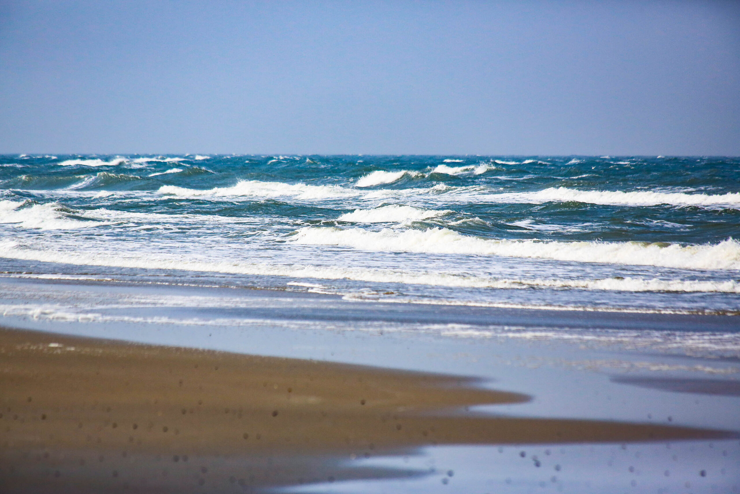 Das Bild zeigt eine Küstenlandschaft, vermutlich an einem sonnigen Tag. Das Hauptaugenmerk liegt auf dem Meer, dessen Wellen an den Sandstrand rollen. Der Sand ist feinkörnig und hellbraun. Der Himmel ist klar und blau, was auf gutes Wetter hindeutet. Die Szene vermittelt ein Gefühl von Ruhe und Weite.