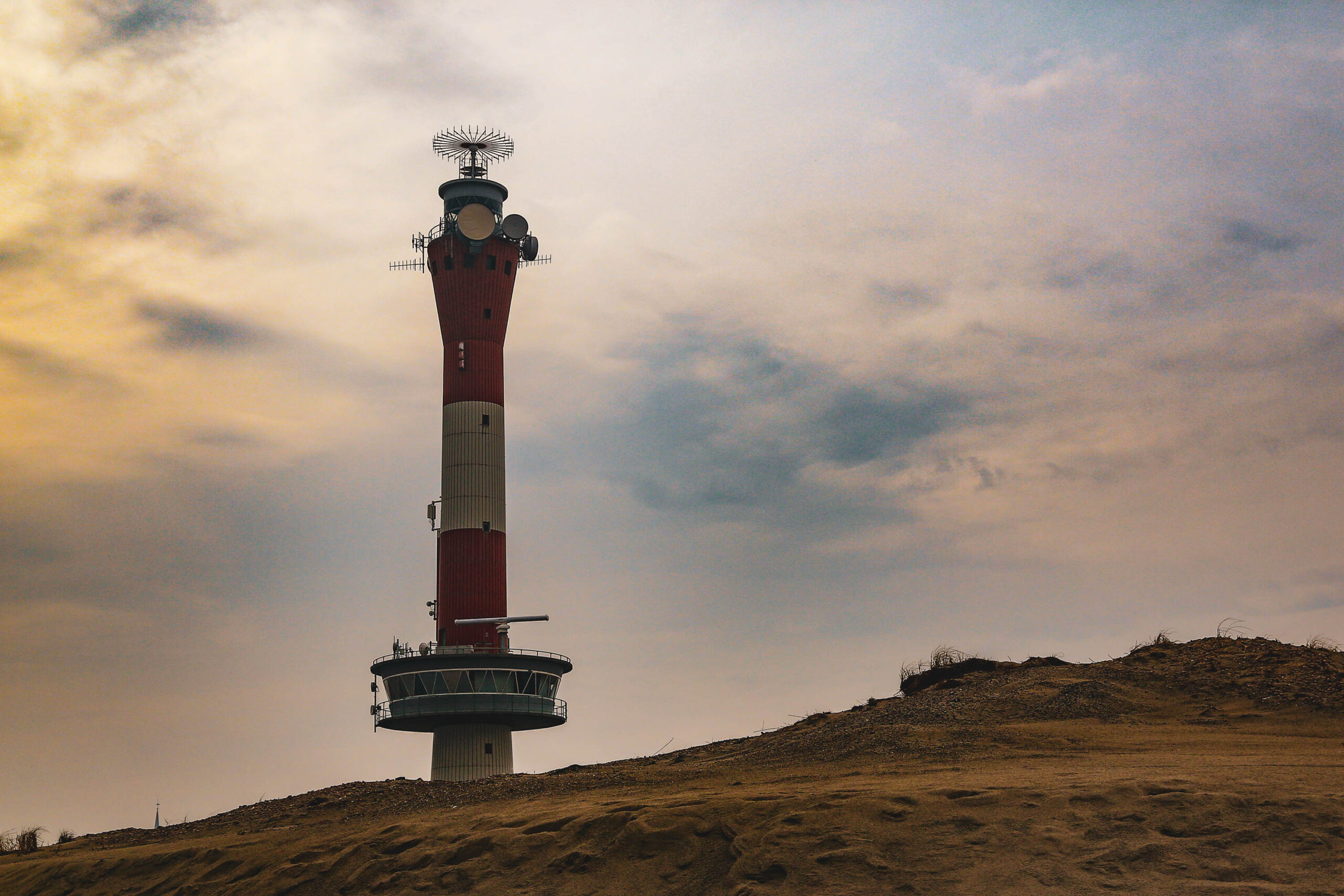 Das Foto zeigt den Leuchtturm von Wangerooge, aufgenommen im April 2018. Der Leuchtturm ist das dominierende Element des Bildes und ragt hoch in den bewölkten Himmel. Er ist rot-weiß gestreift und besitzt eine komplexe Anordnung von Antennen und Plattformen an der Spitze. Der Leuchtturm steht auf einem sandigen Hügel, der mit niedrigem Gras und einigen Büschen bewachsen ist. Der Himmel ist dunkel und bewölkt, was eine düstere und melancholische Stimmung erzeugt. Die Perspektive ist von unten, was die Höhe des Leuchtturms betont. Das Bild fängt die raue Schönheit der Nordseeinsel Wangerooge ein.