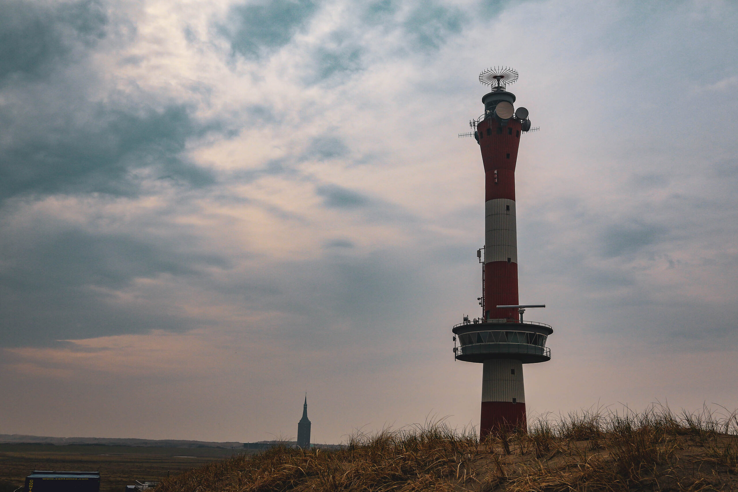 Das Bild zeigt einen hohen Leuchtturm auf der Insel Wangerooge. Der Leuchtturm ist in rote und weiße Streifen gemustert und hat eine Plattform mit Antennen auf dem Kopf. Im Hintergrund sind Dünen zu sehen, die mit trockenem Gras bewachsen sind. Der Himmel ist dunkel und bewölkt, was eine düstere Stimmung erzeugt. Ein weiterer, kleinerer Turm ist im Hintergrund zu erkennen. Das Bild wurde im April 2018 aufgenommen.
