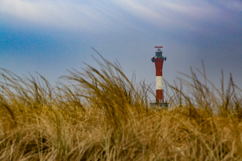 Das Bild zeigt einen Leuchtturm, der sich aus dichtem, hohem Gras erhebt. Der Himmel ist bedeckt und erstreckt sich in einem bläulichen Farbton. Das Gras dominiert den unteren Teil des Bildes und verleiht der Szene eine organische und natürliche Atmosphäre. Der Leuchtturm ist in Weiß und Rot gestrichen und ragt deutlich in den Himmel. Die Komposition betont die Isolation und die Weite der Landschaft.