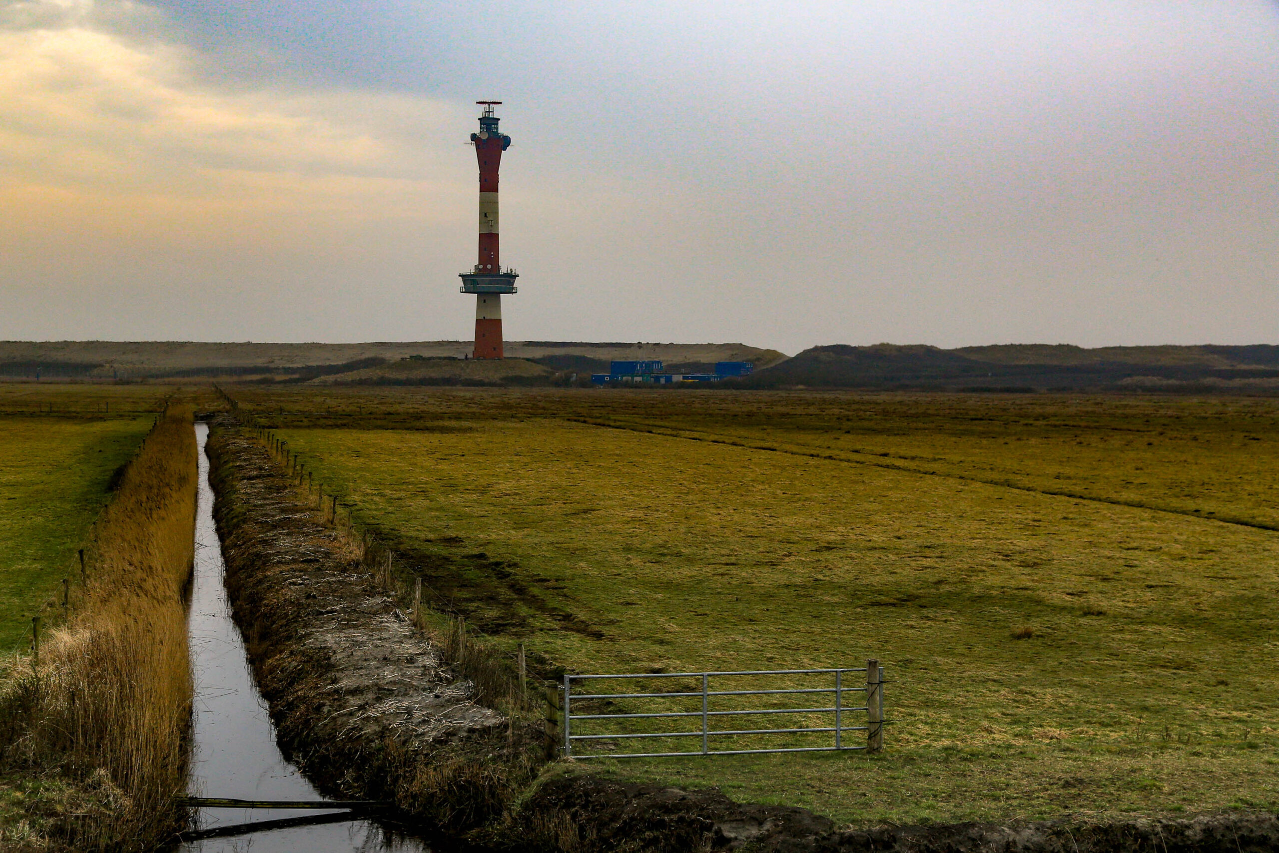Die Aufnahme zeigt eine typische Landschaft der ostfriesischen Insel Wangerooge. Im Vordergrund fließt ein schmaler, dunkler Graben, dessen Ufer von dichtem Gras bewachsen sind. Ein Metallzaun versperrt den Zugang zum Graben. Im Hintergrund erhebt sich ein Leuchtturm, der die Silhouette der Insel dominiert. Der Himmel ist bedeckt und erzeugt eine gedämpfte, melancholische Stimmung. Die Farben sind gedämpft, was die Atmosphäre der Insel unterstreicht.