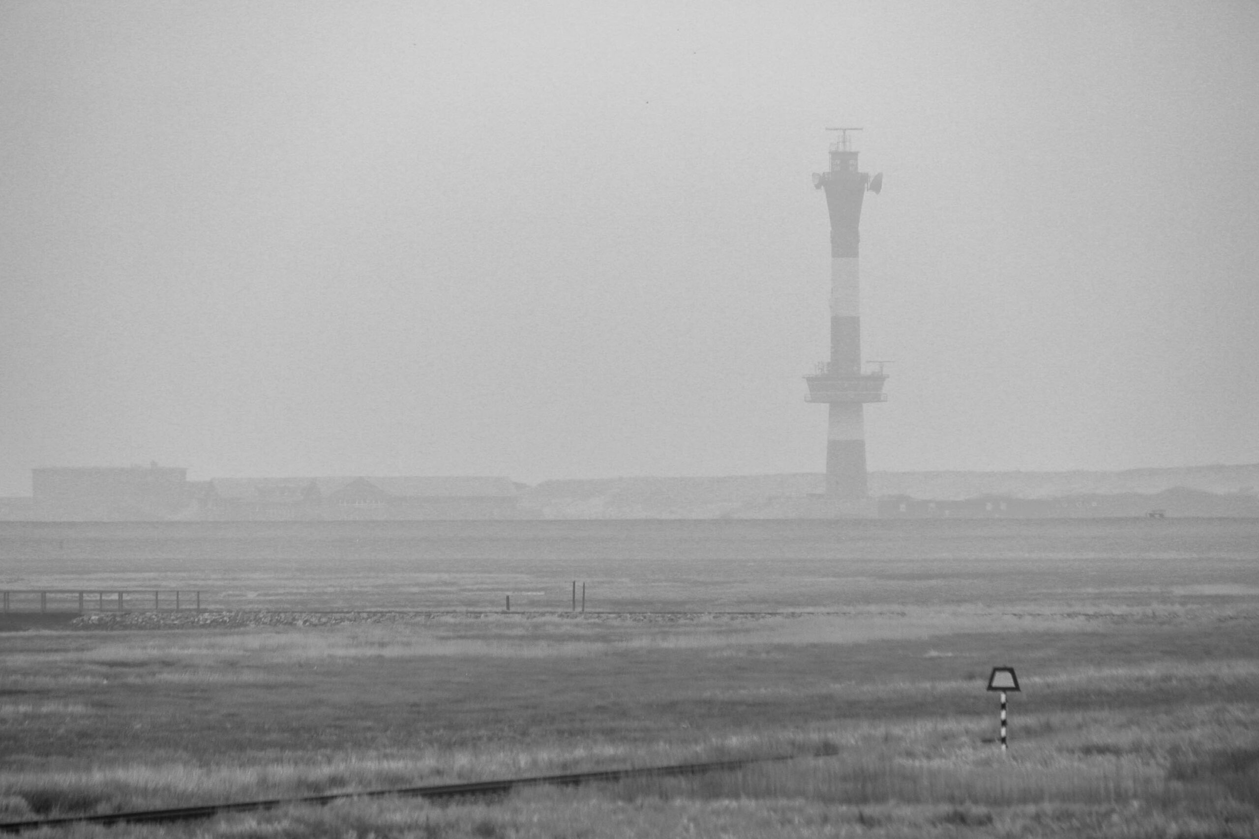 Das Foto zeigt eine neblige Landschaft auf der Insel Wangerooge. Im Vordergrund erstreckt sich eine flache, grasbewachsene Fläche, die in den Horizont übergeht. Ein Leuchtturm ragt aus der nebligen Szenerie auf, sein Turm ist deutlich erkennbar, während die Basis im Nebel verschwimmt. Im Hintergrund sind Gebäude angedeutet, die ebenfalls vom Nebel verhüllt sind. Ein einzelner, schwarzer Wegweiser steht isoliert im Vordergrund. Die gesamte Szene ist in Schwarzweiß gehalten, was die düstere und neblige Atmosphäre verstärkt.